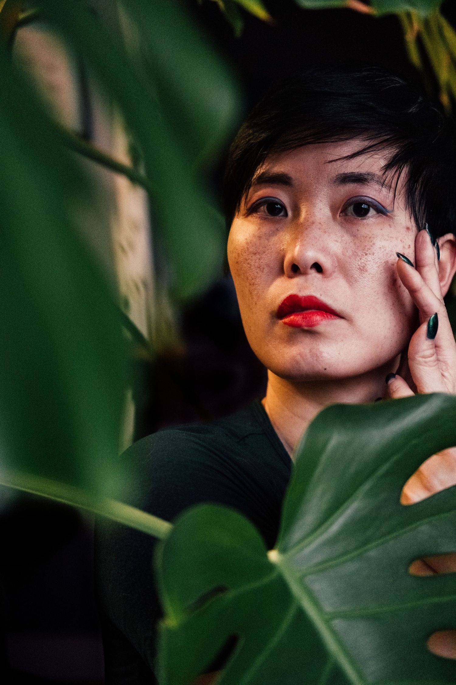 Artistic portrait through green leaves showing dramatic lighting and red lipstick against a dark background.