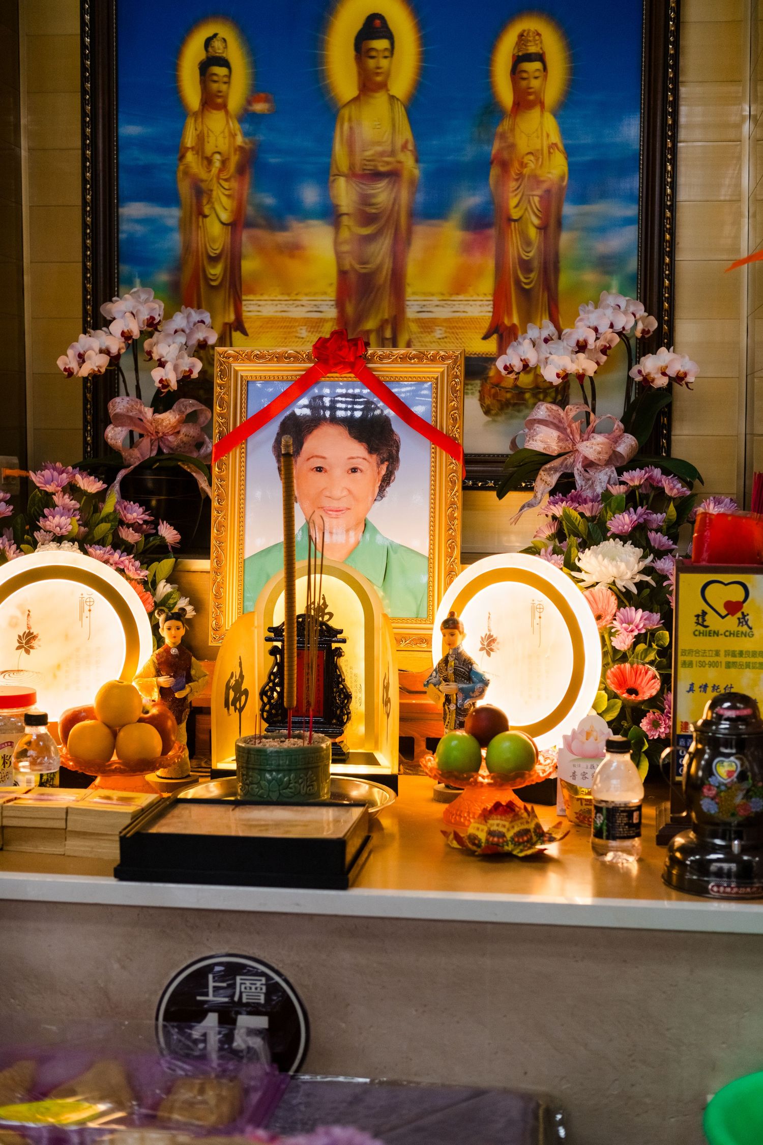 Buddhist altar display with golden statues, pink orchids, and memorial photographs in a temple setting.