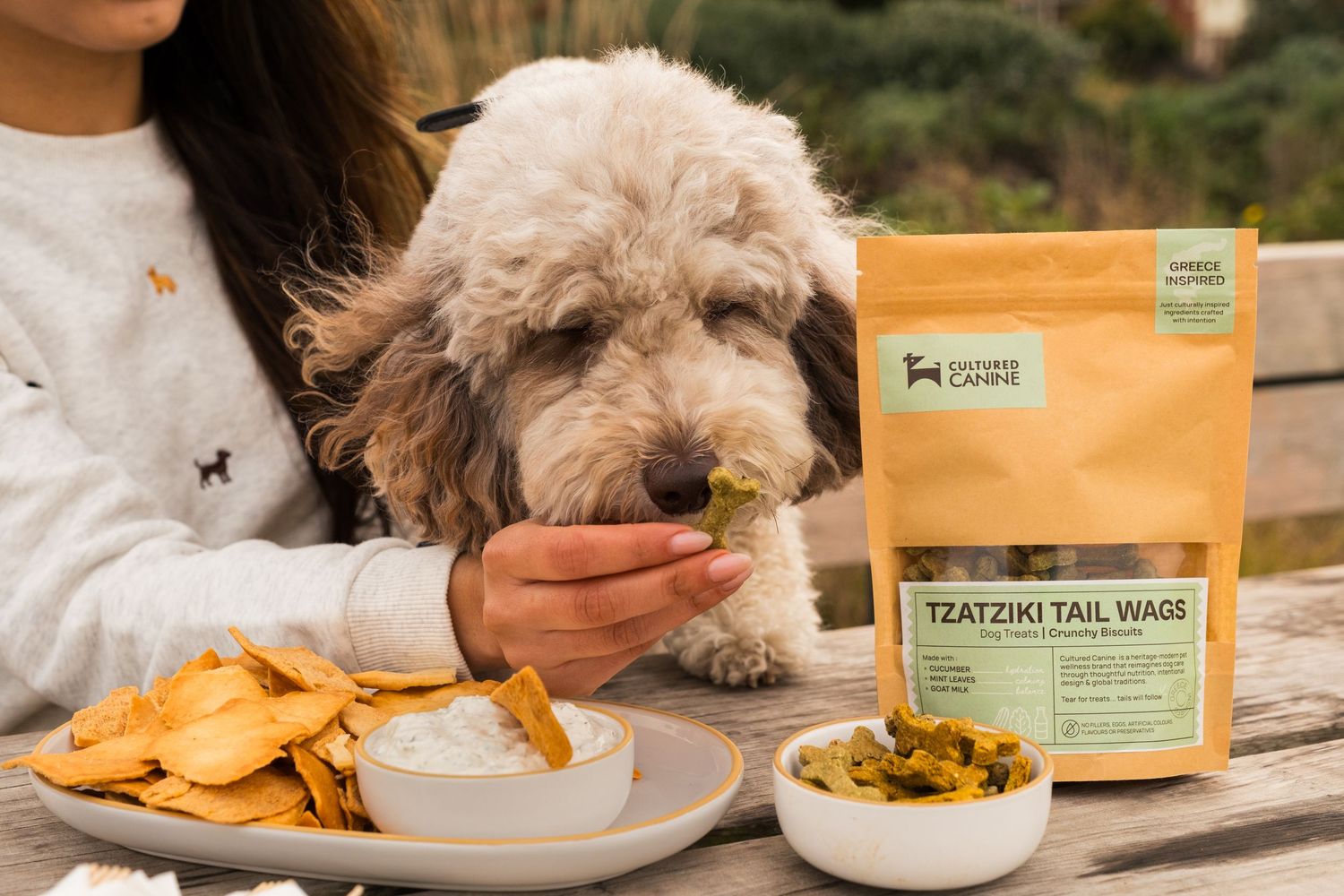 A fluffy dog sniffs a treat held by a woman beside Tzatziki Tail Wags dog treats bag on a wooden table.