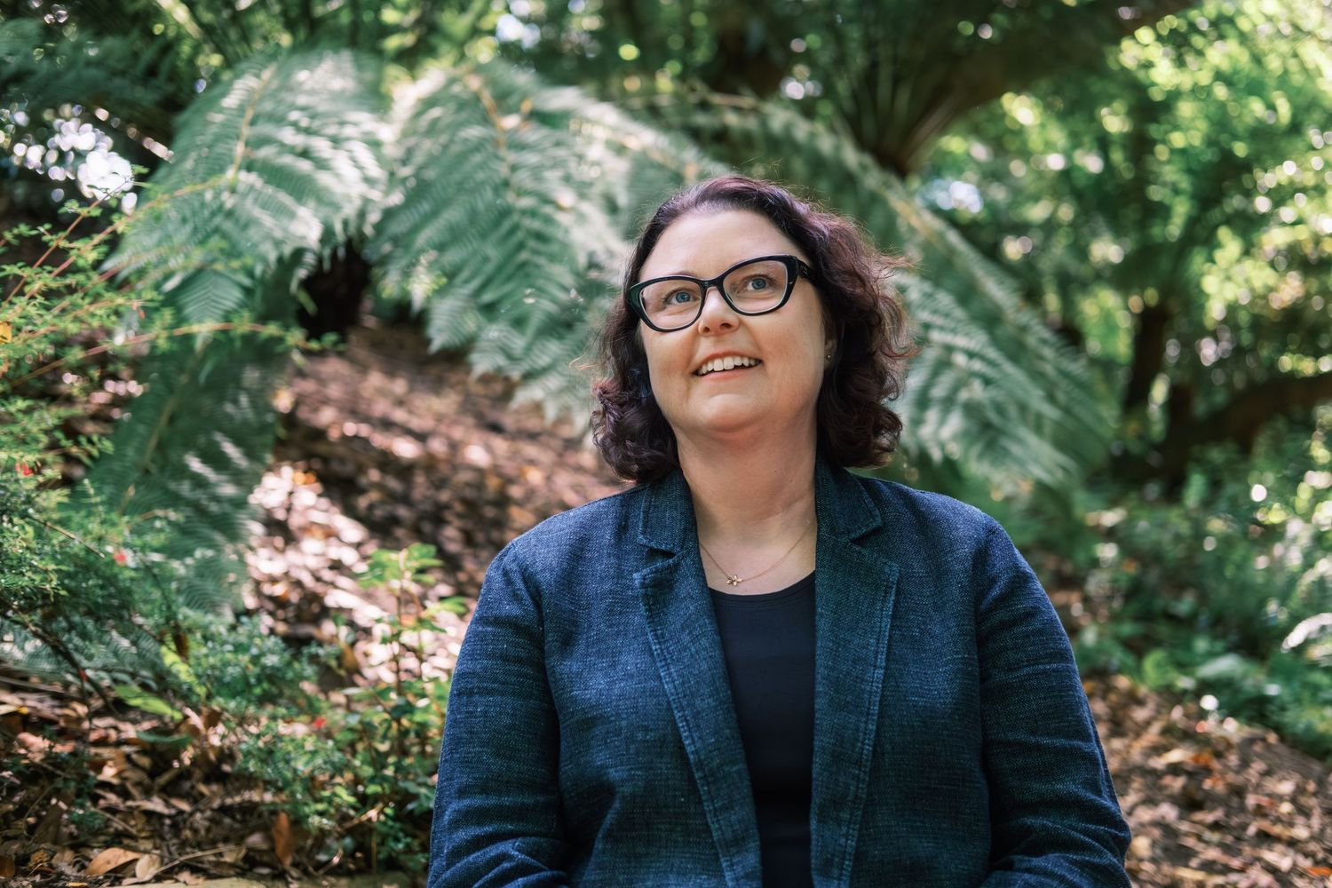 A person in glasses and a blue blazer sits outdoors surrounded by lush greenery in a natural setting.