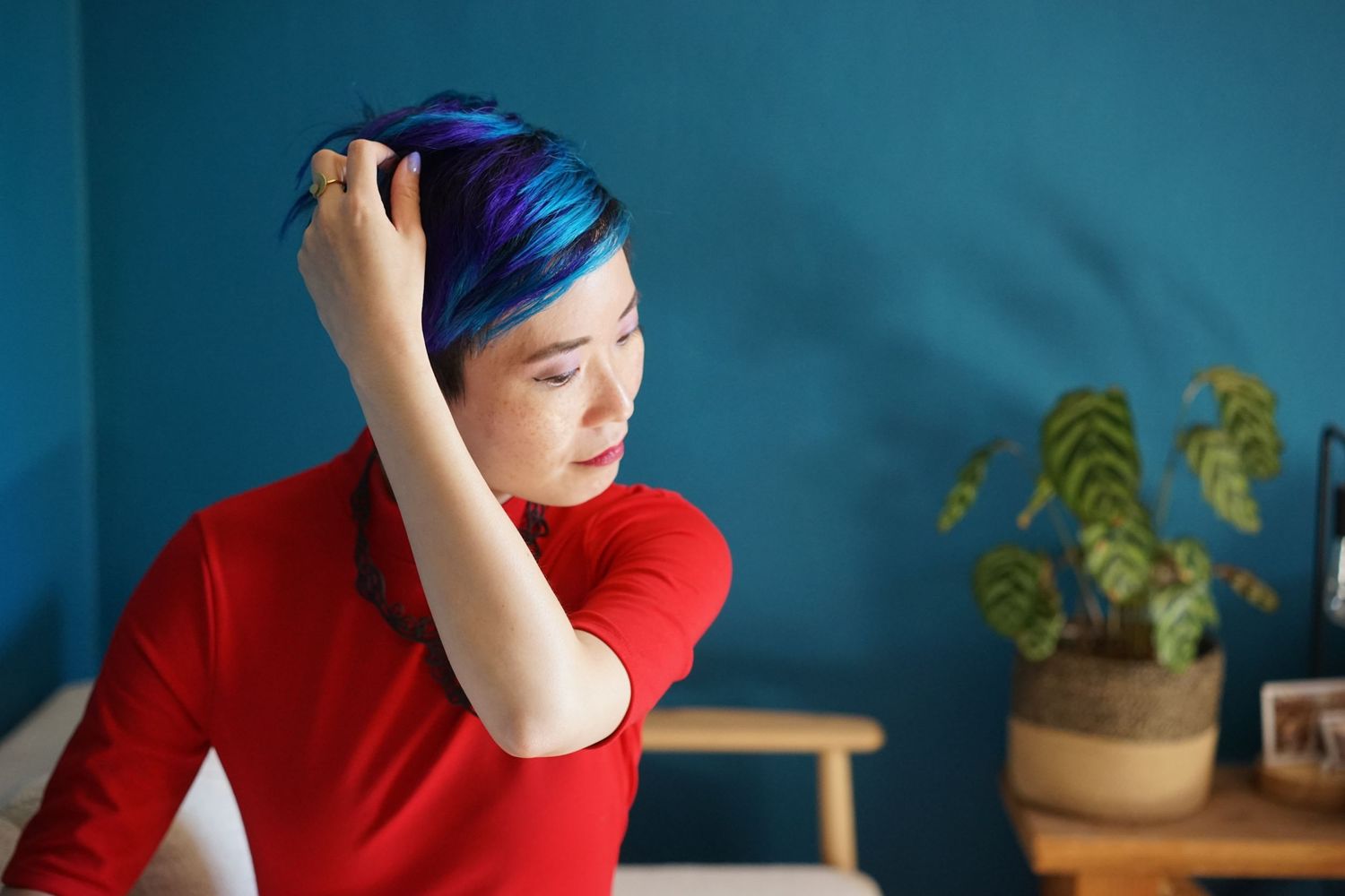 Person with vibrant blue hair in red shirt against teal wall with plant.