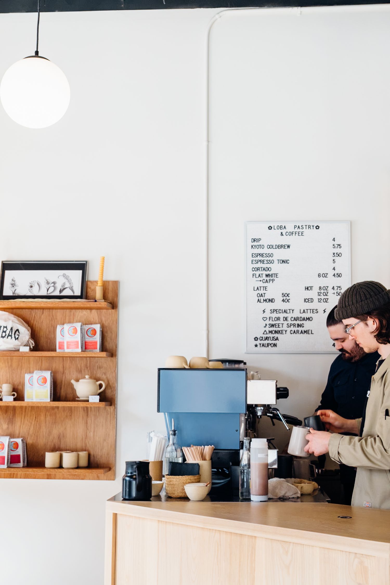 A barista works at a coffee counter with a blue espresso machine and wooden shelving displaying coffee accessories and decor.
