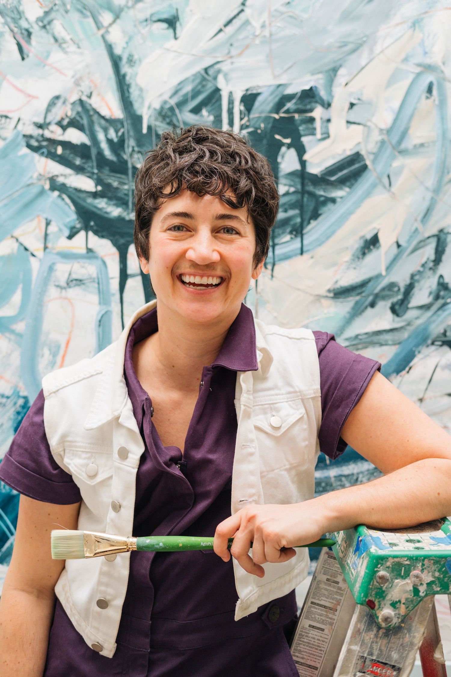 Artist in purple shirt and white vest holds paintbrush against abstract blue background.