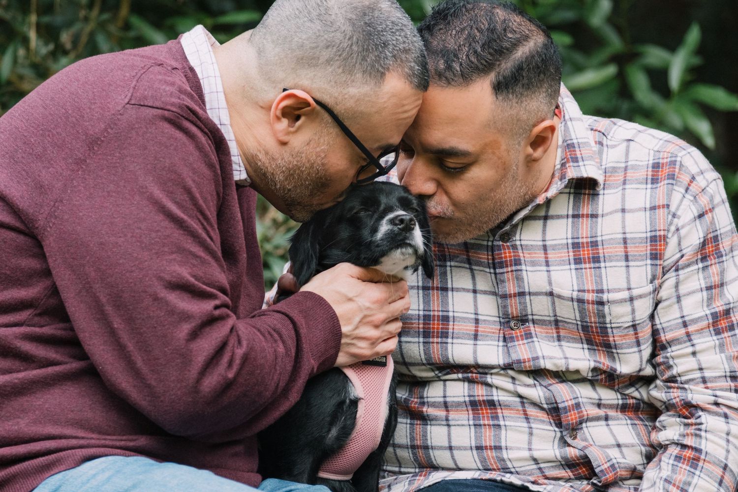 Two men sharing a tender moment kissing their small black dog outdoors among green foliage.