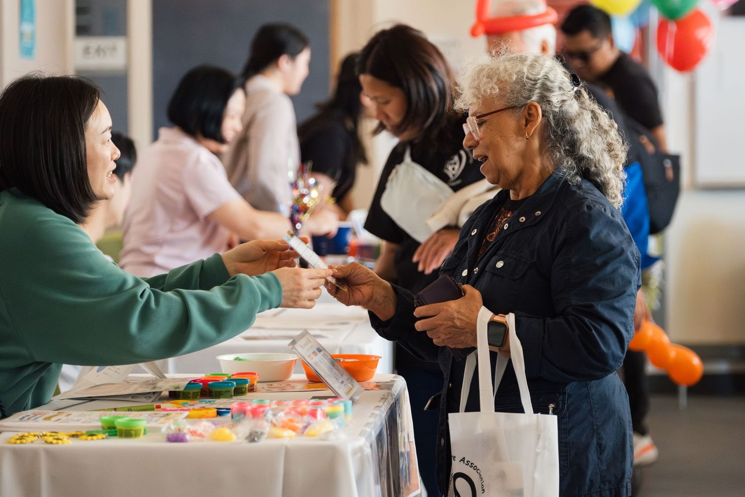 People networking at a community fair event, exchanging items at a table with colorful decorations and balloons in background.