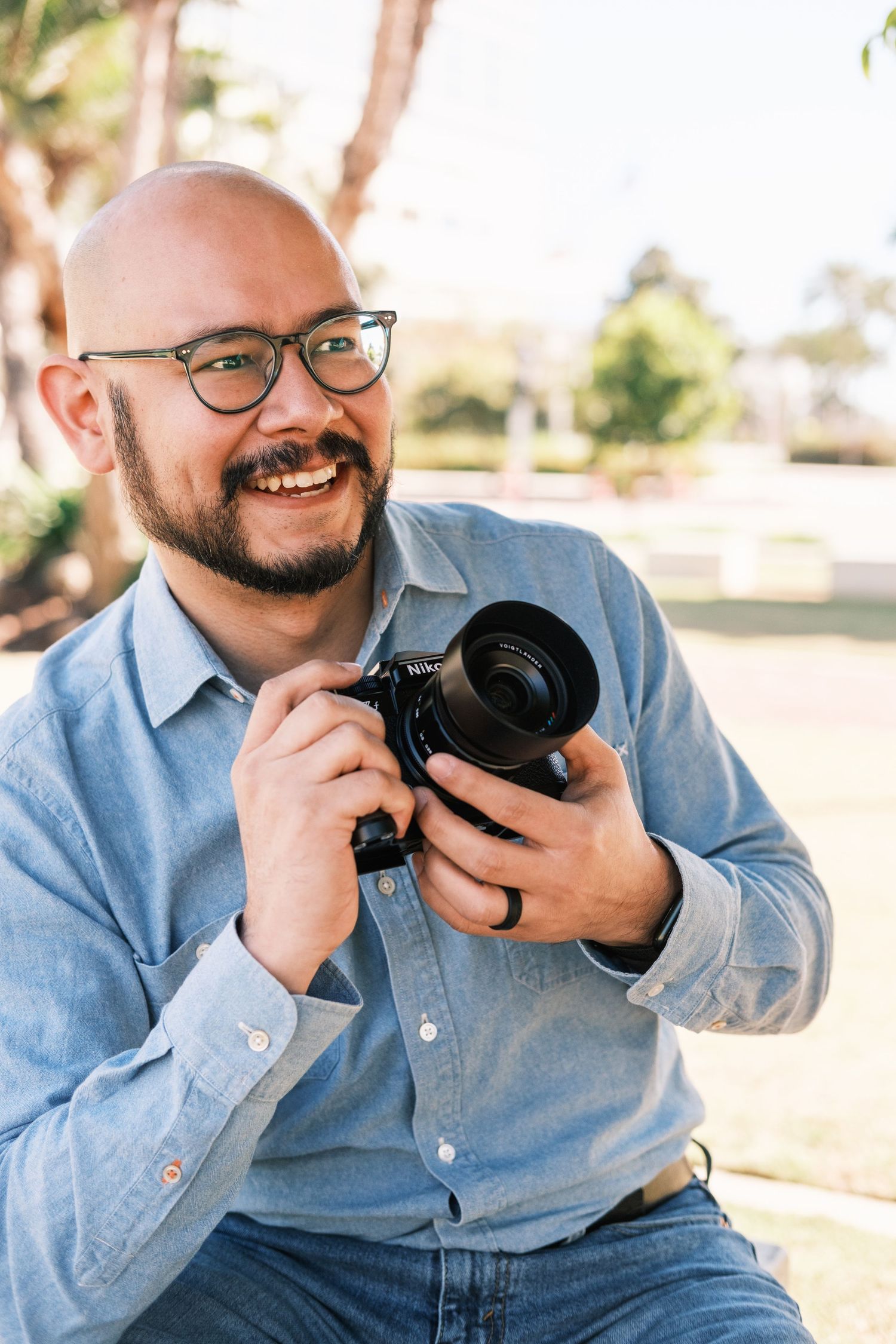Smiling bald man with glasses holding a camera outdoors, wearing a blue shirt in a sunny park setting.