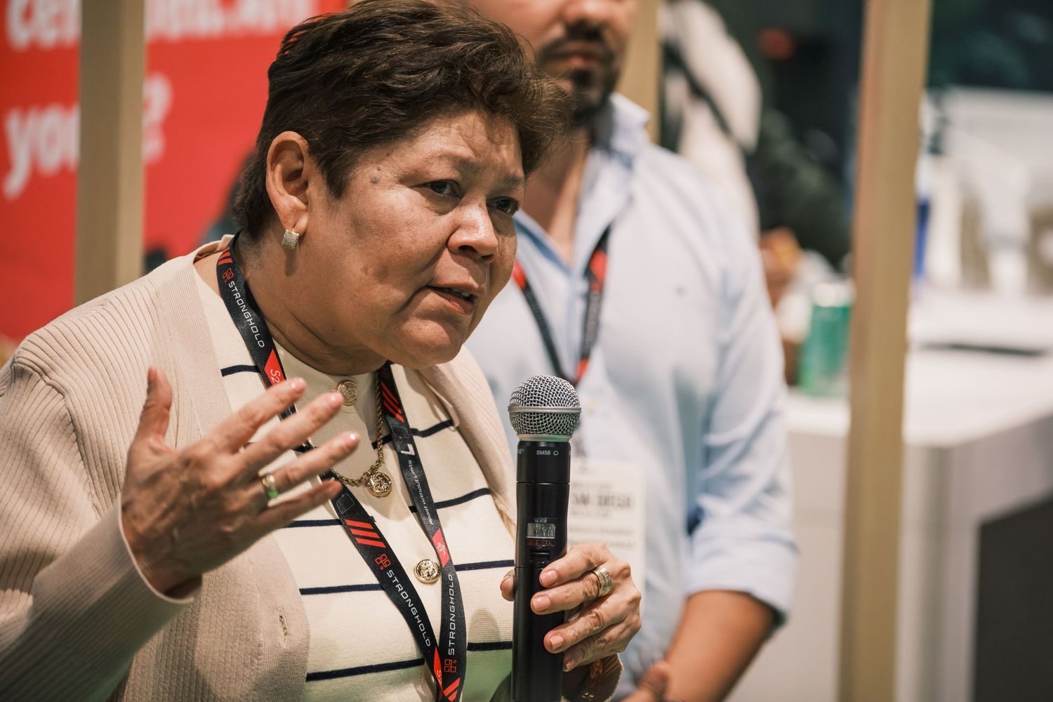 Woman holding microphone speaking at conference event with lanyard badge, gesturing expressively indoors.