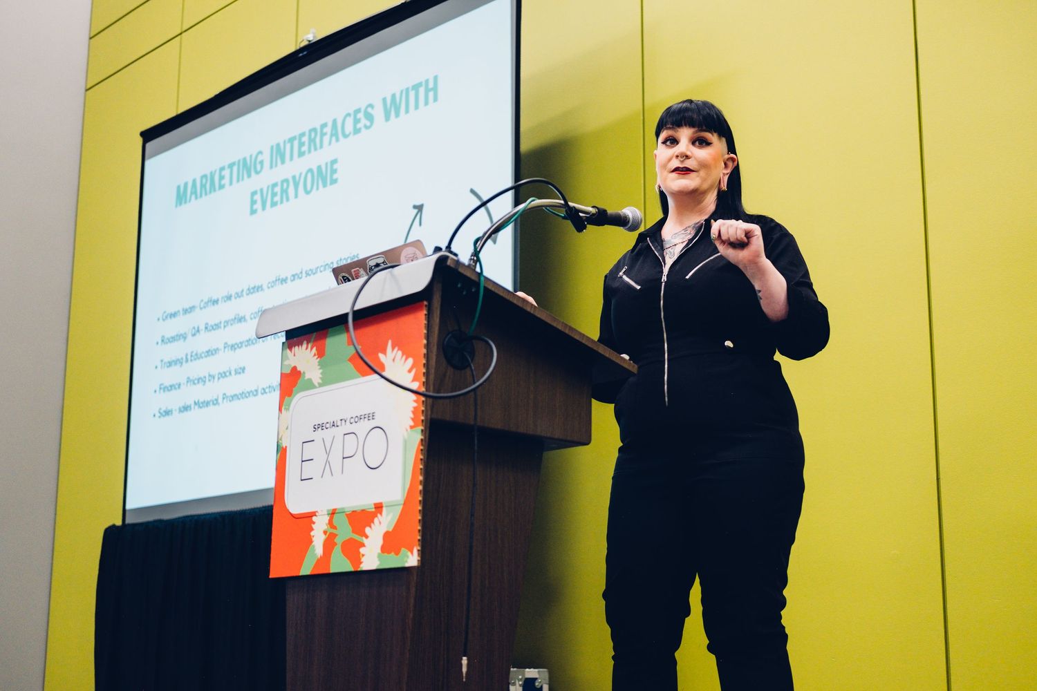 A presenter in black attire gestures while speaking at a podium during a conference presentation.