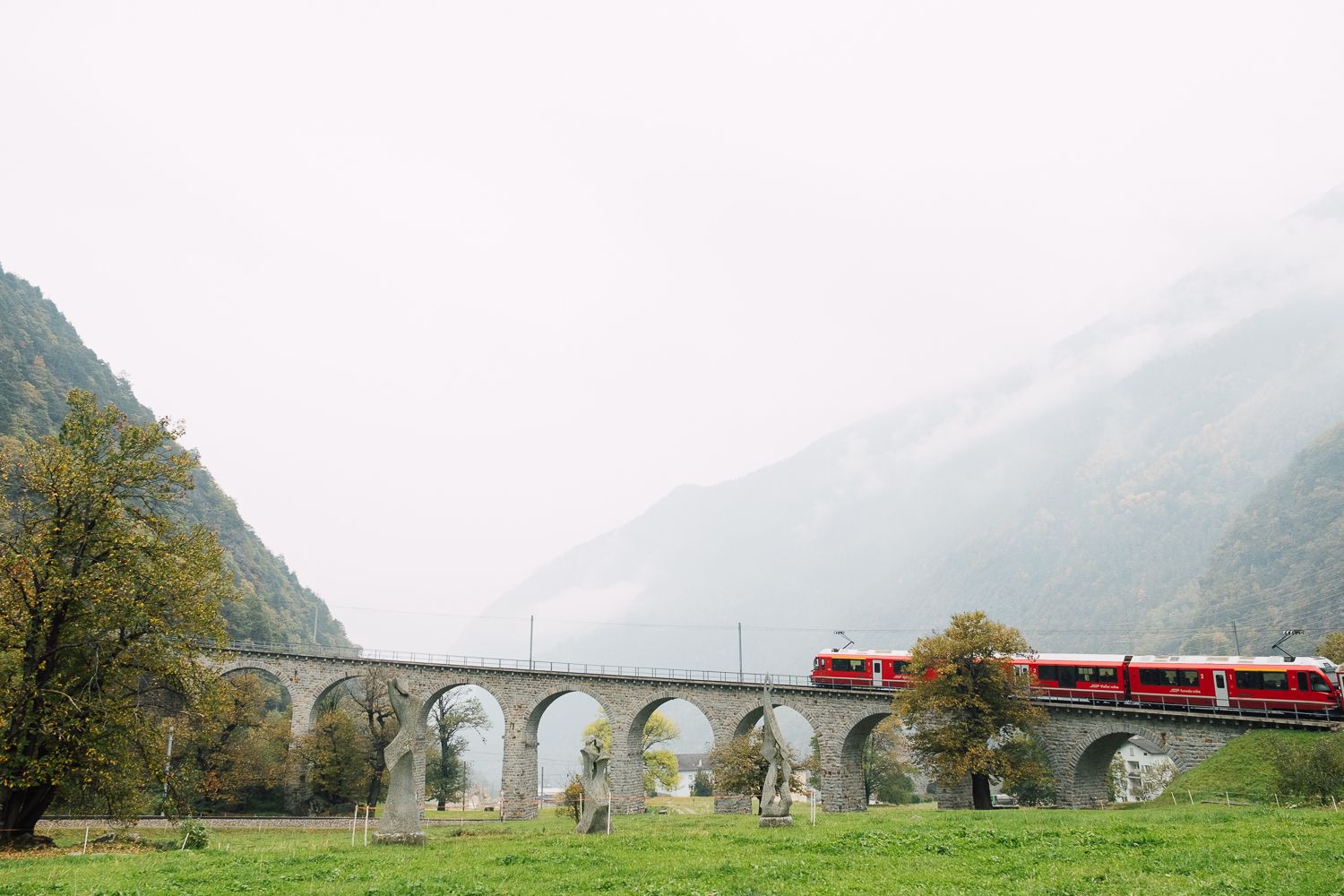 A red train crosses a curved stone viaduct bridge through misty mountains in a scenic landscape setting.