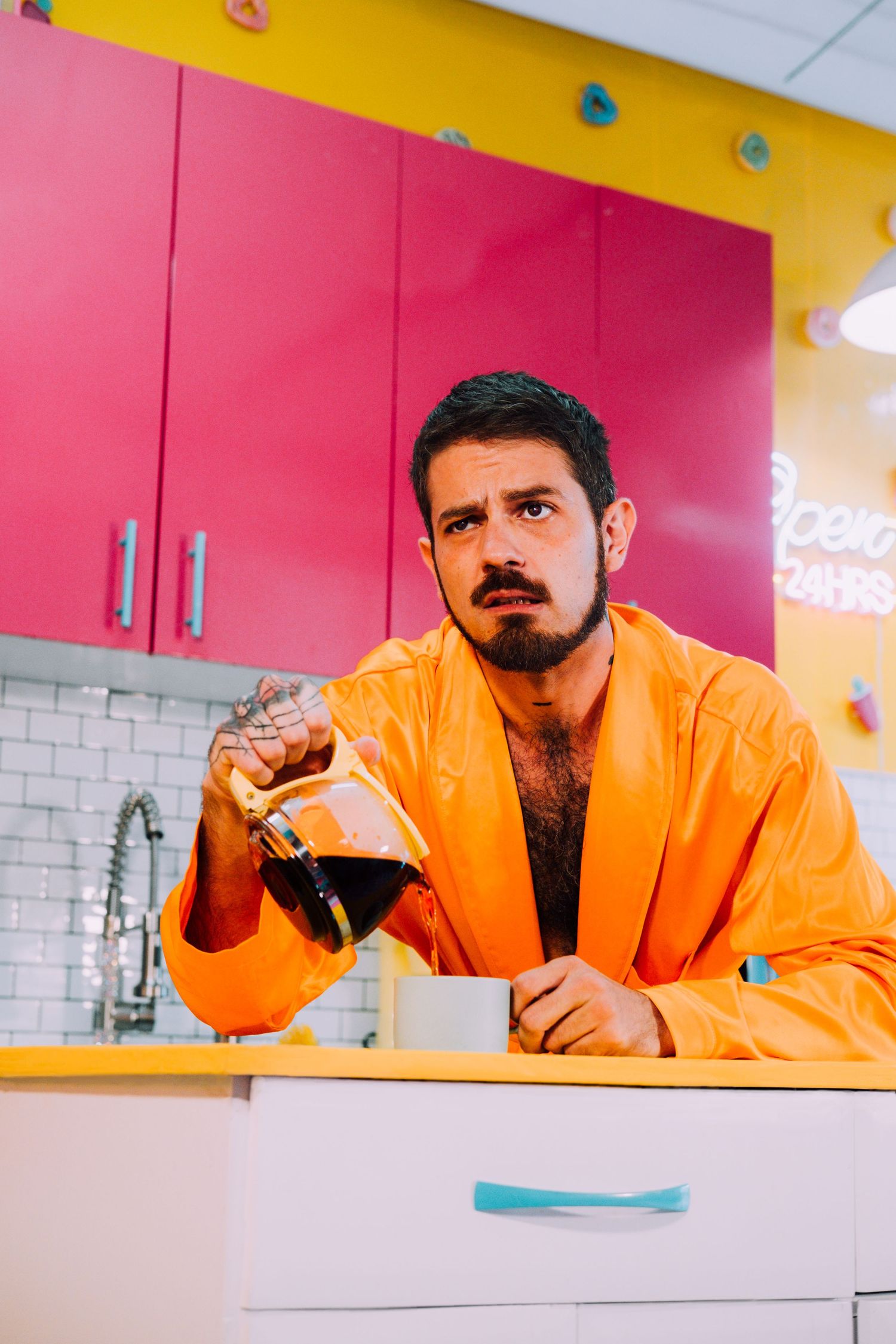 Person in bright orange robe holds coffee while sitting at counter with pink and yellow kitchen backdrop.