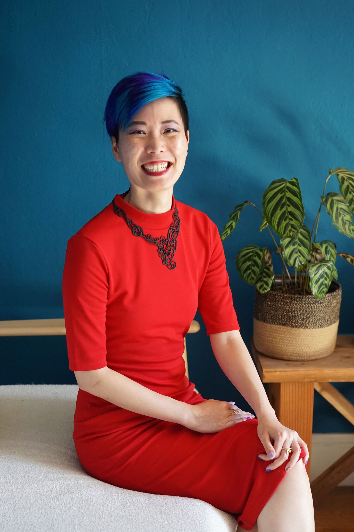 A person in a red dress and statement necklace sits smiling against a blue wall with a potted plant.