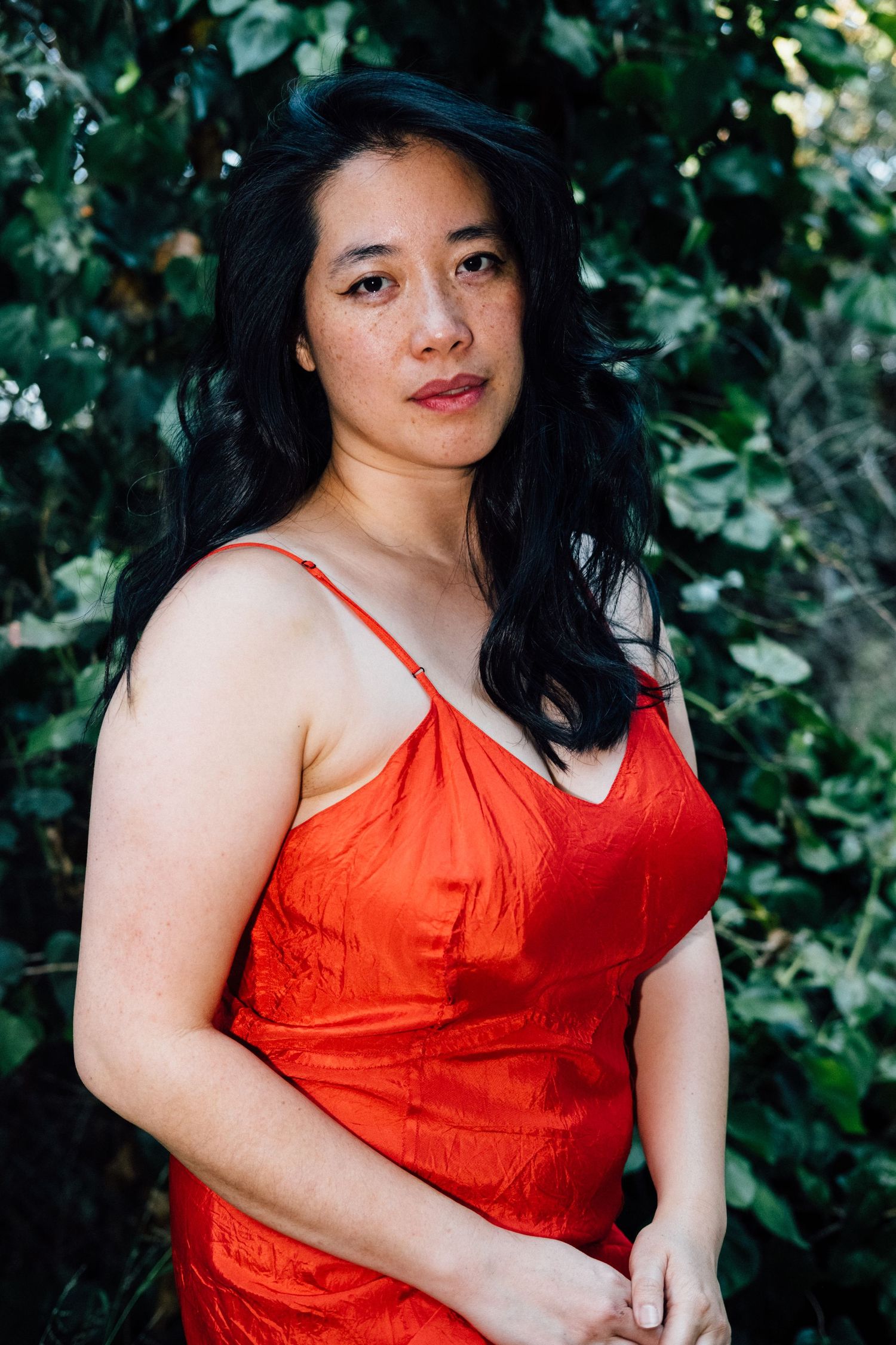 A person in a bright red sleeveless dress poses against a backdrop of lush green foliage in an outdoor setting.