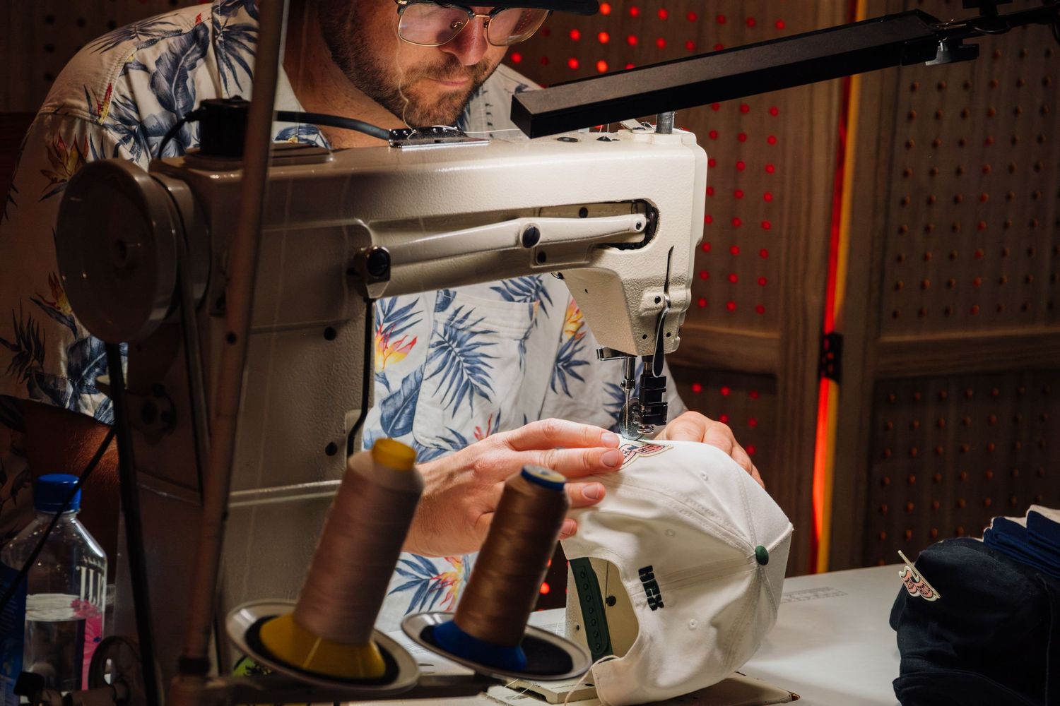A tailor in a floral shirt operates a sewing machine with brown thread spools, carefully stitching white fabric.