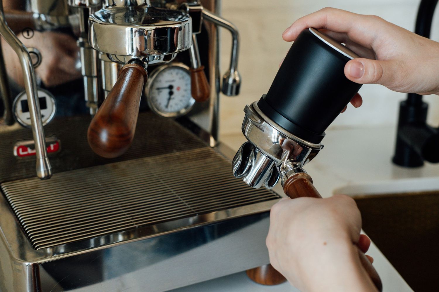 Hands adjusting coffee grounds in a portafilter near an espresso machine.