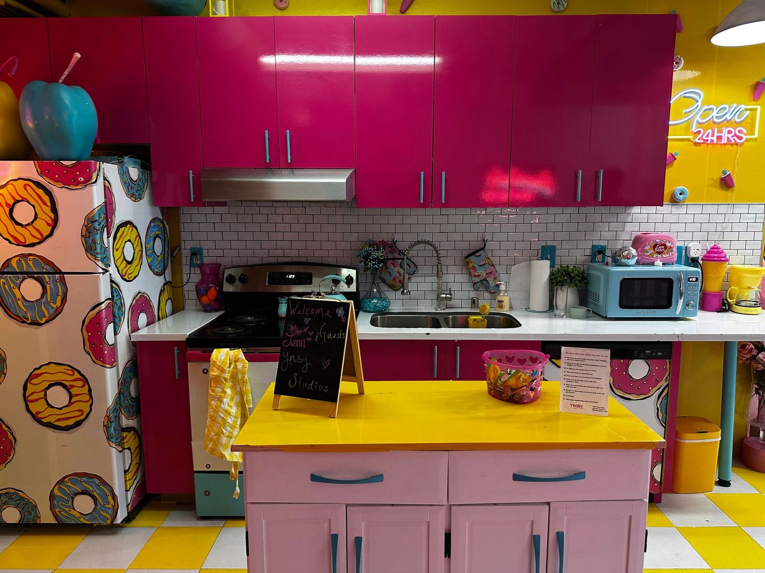 Retro-styled kitchen with bright pink upper cabinets, yellow countertops, donut wallpaper, and colorful decorative elements.
