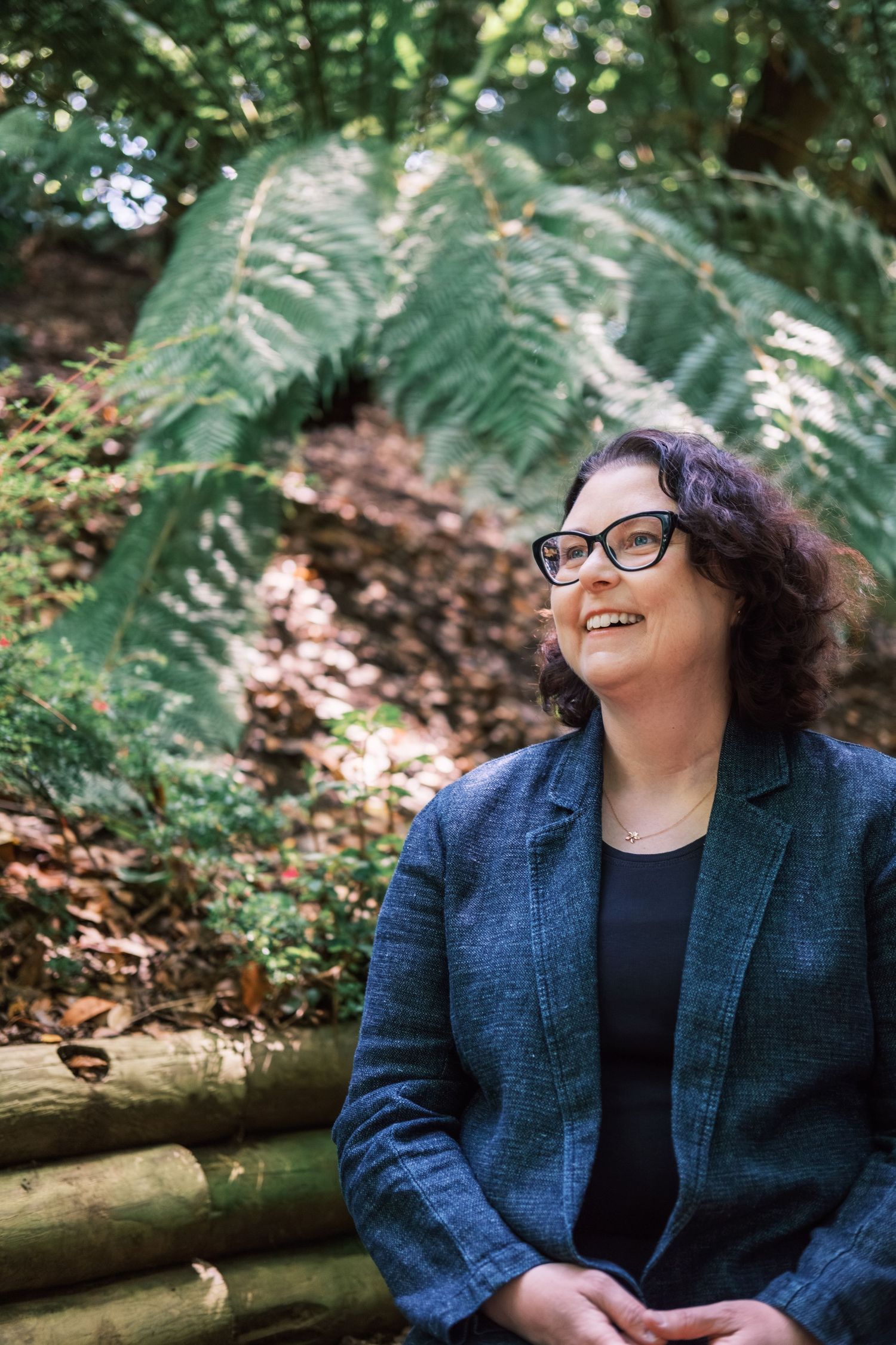 A series of candid outdoor portraits showing someone in a navy blazer and glasses sitting near ferns and foliage.