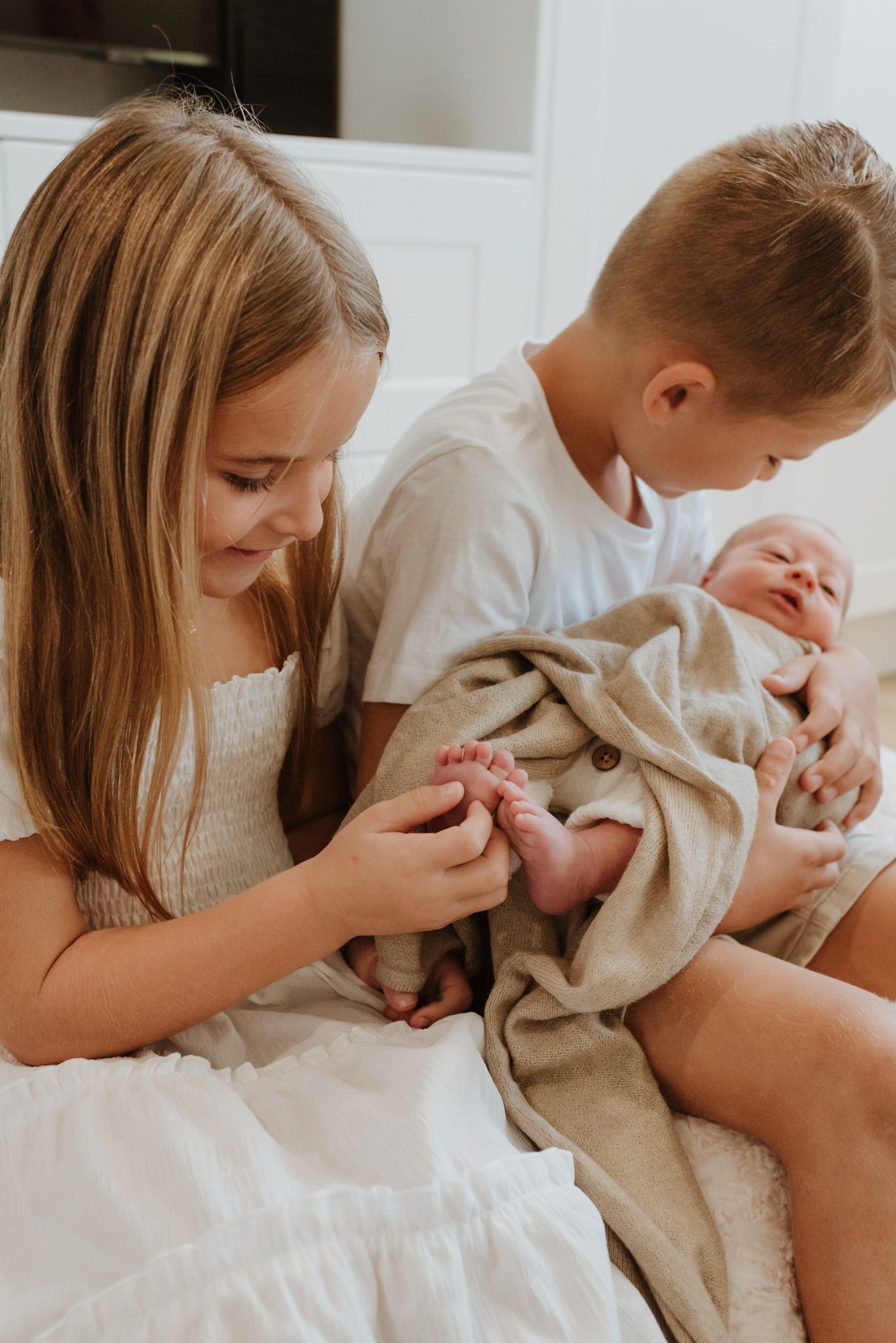 Children in white clothes sitting on bed with newborn baby wrapped in beige blanket.