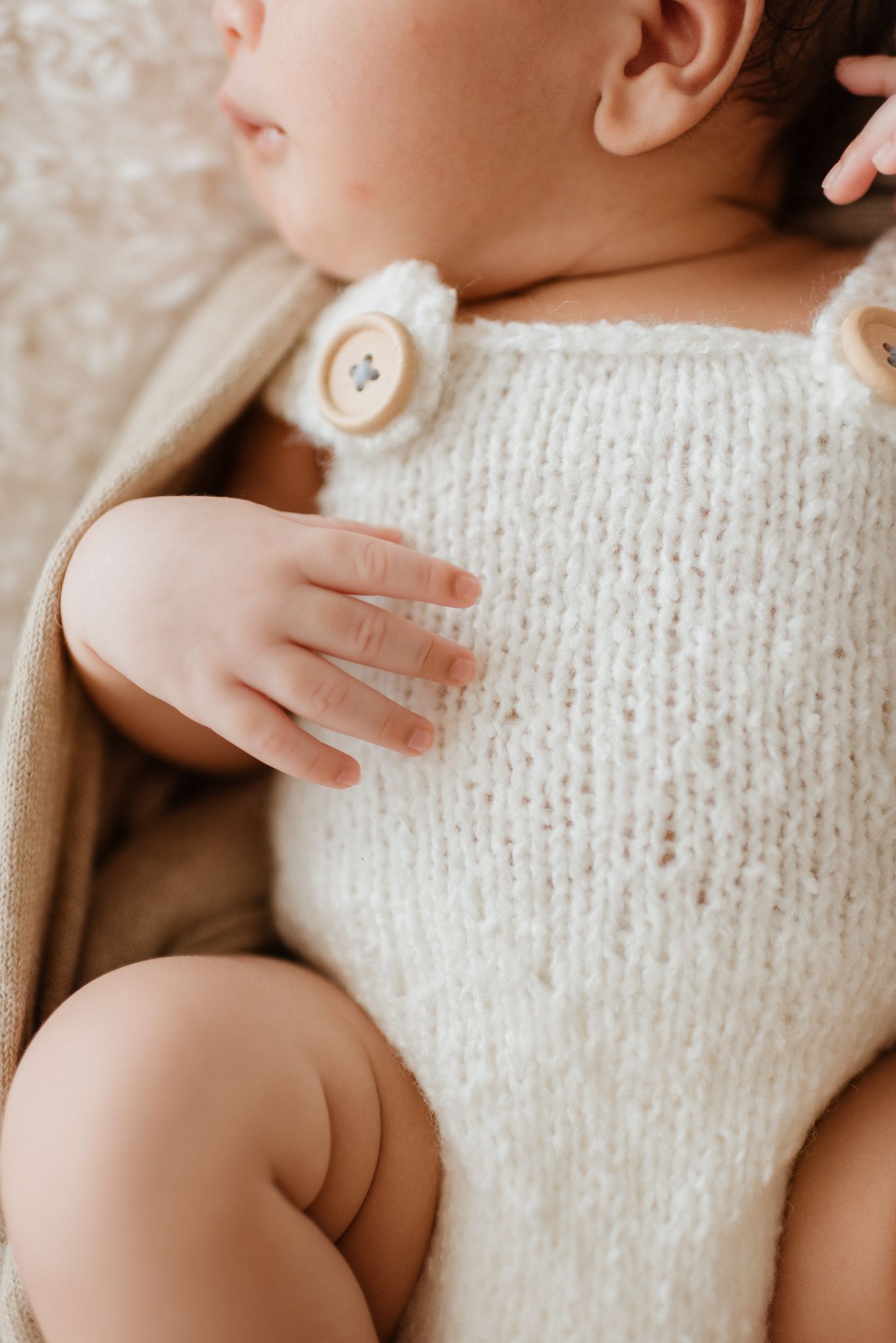 Close-up of tiny infant hand against cream knit sweater with button detail in warm natural lighting.