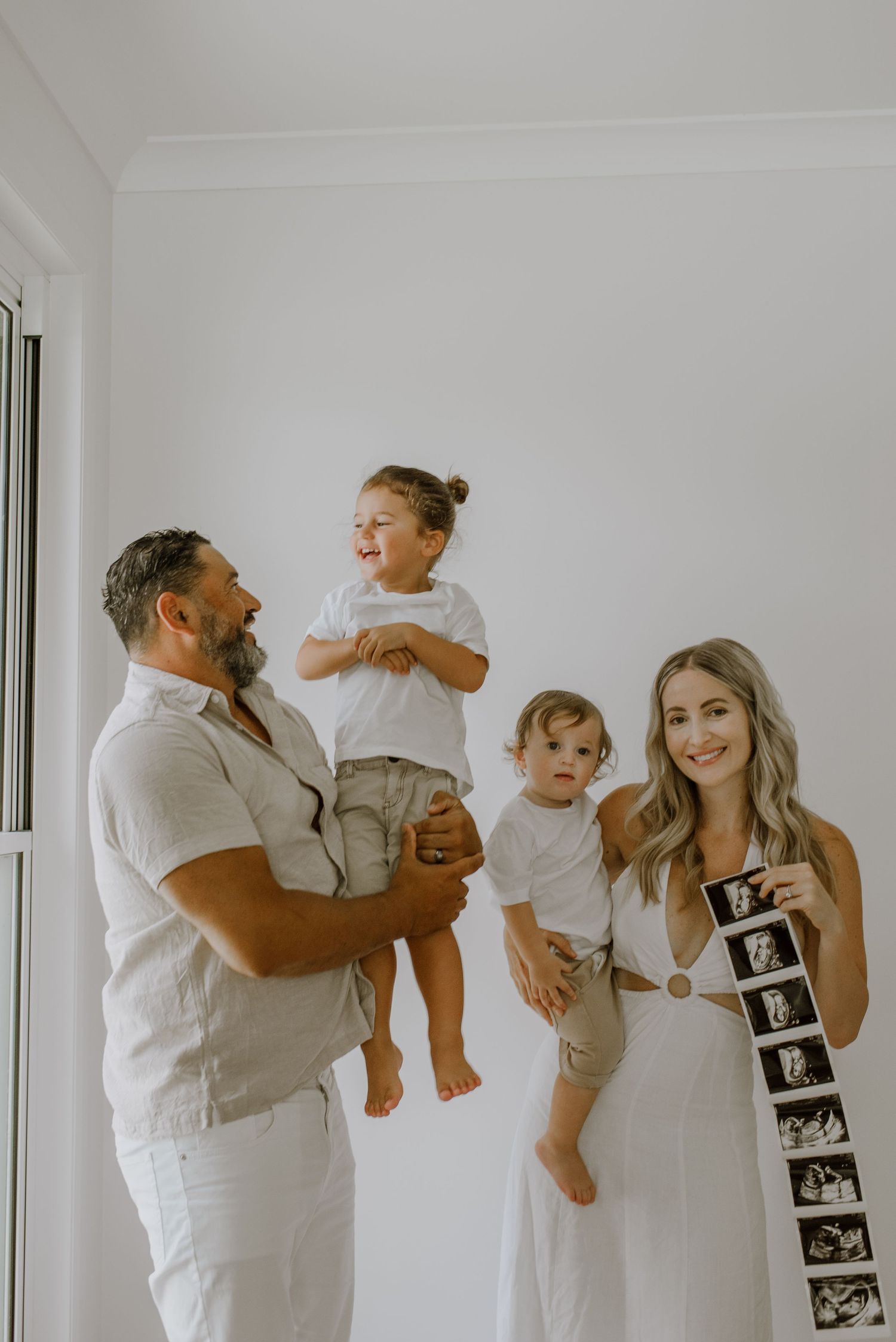 A family in white clothing poses together in a minimalist setting with ultrasound photos displayed on the wall.
