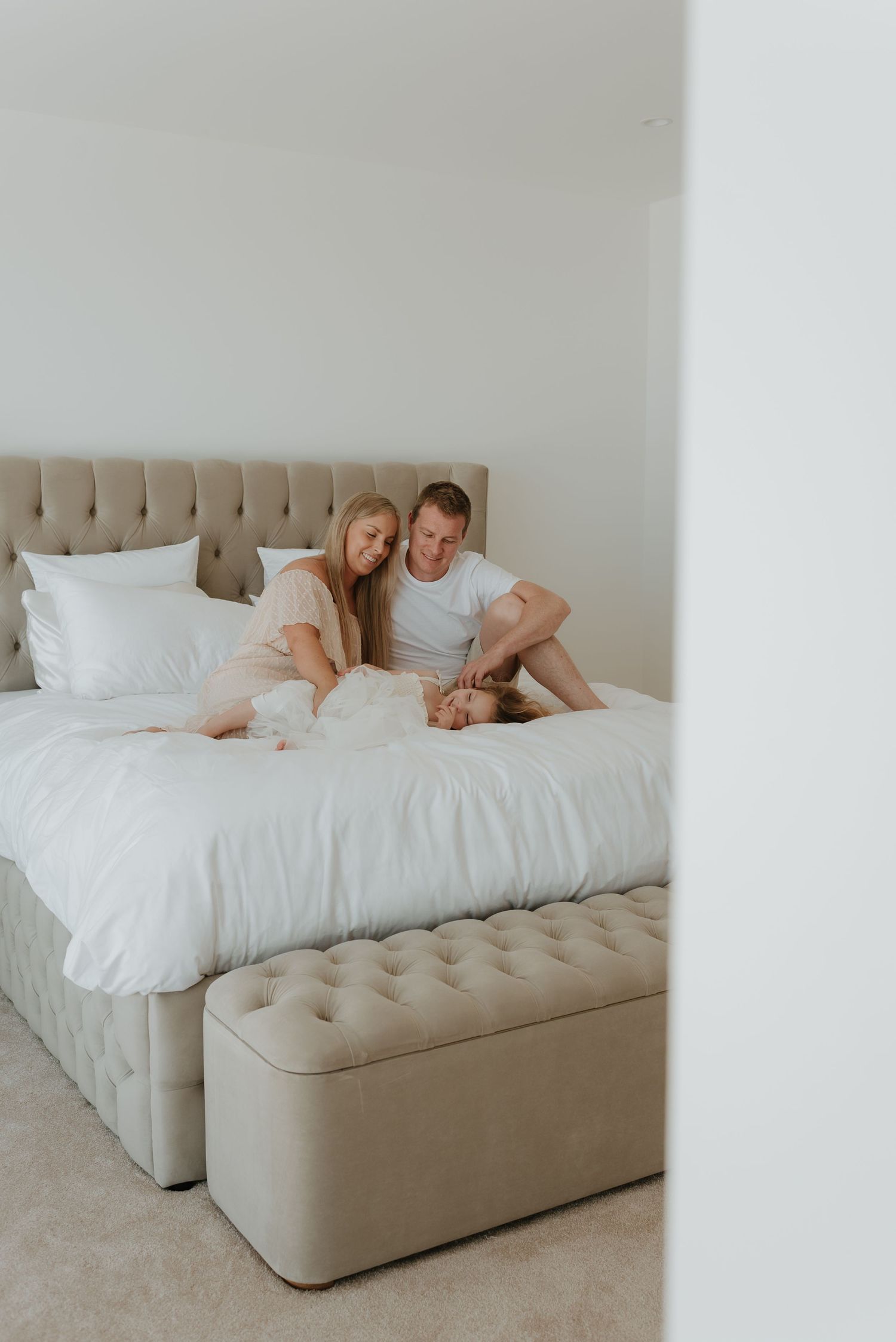 A happy couple relaxing on a large tufted bed with white bedding in a bright, minimalist bedroom.