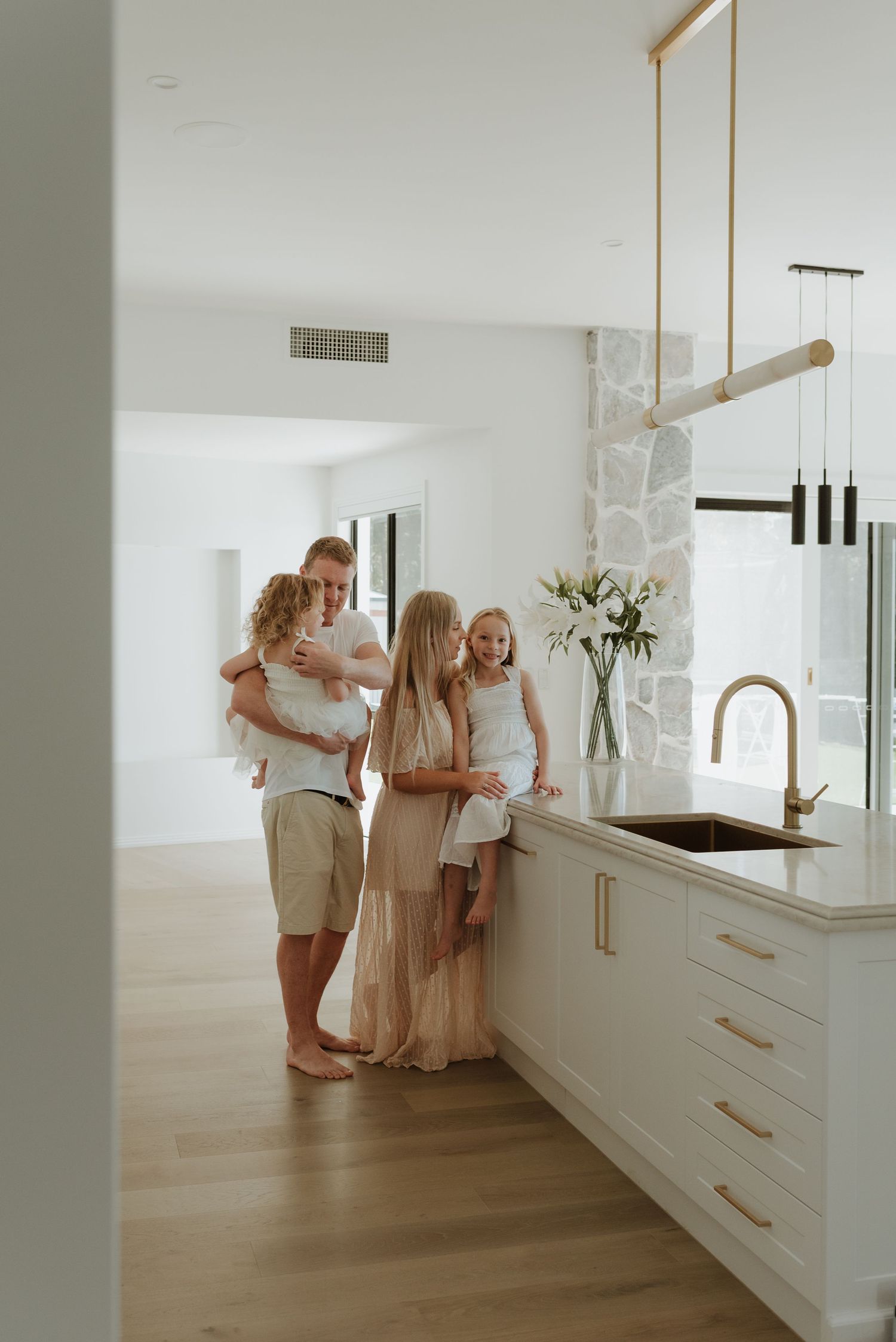 People in white clothing interact in a bright, modern kitchen space with light wood floors and white cabinetry.