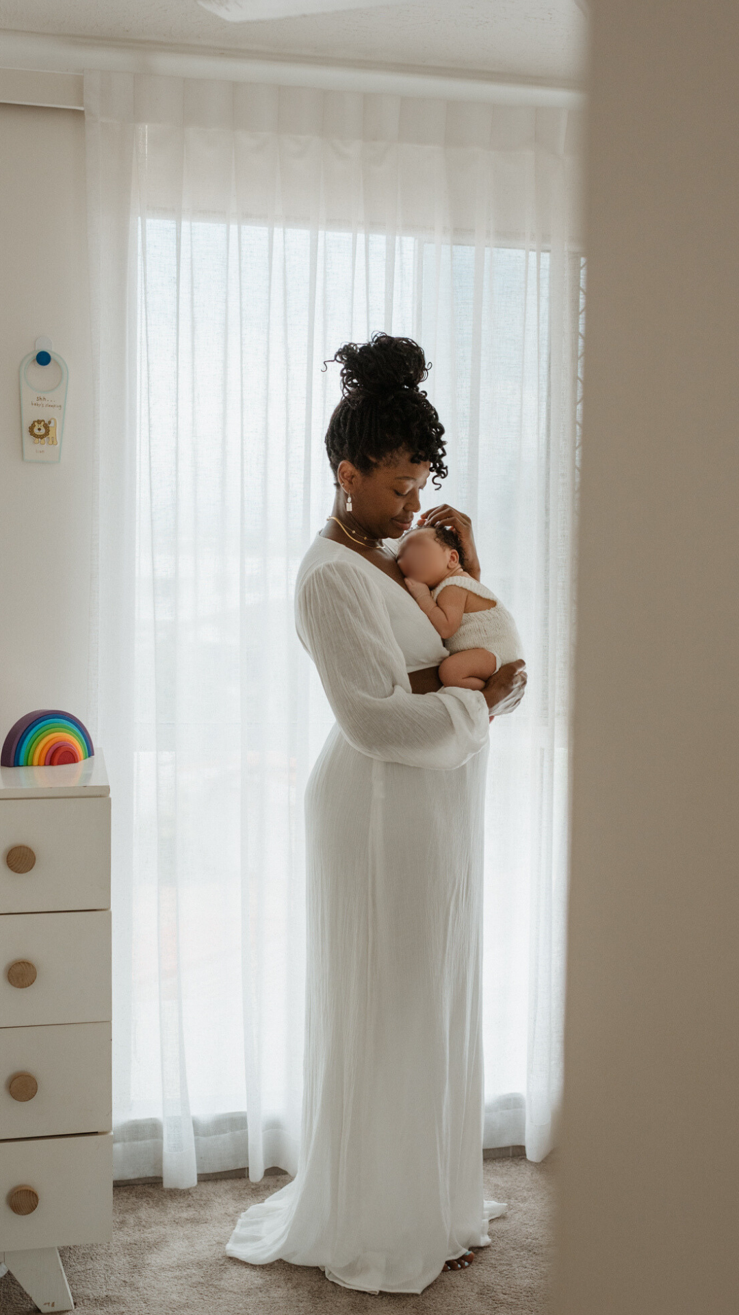 A mother in a flowing white dress holds her newborn baby near a window with sheer curtains in a bright nursery.