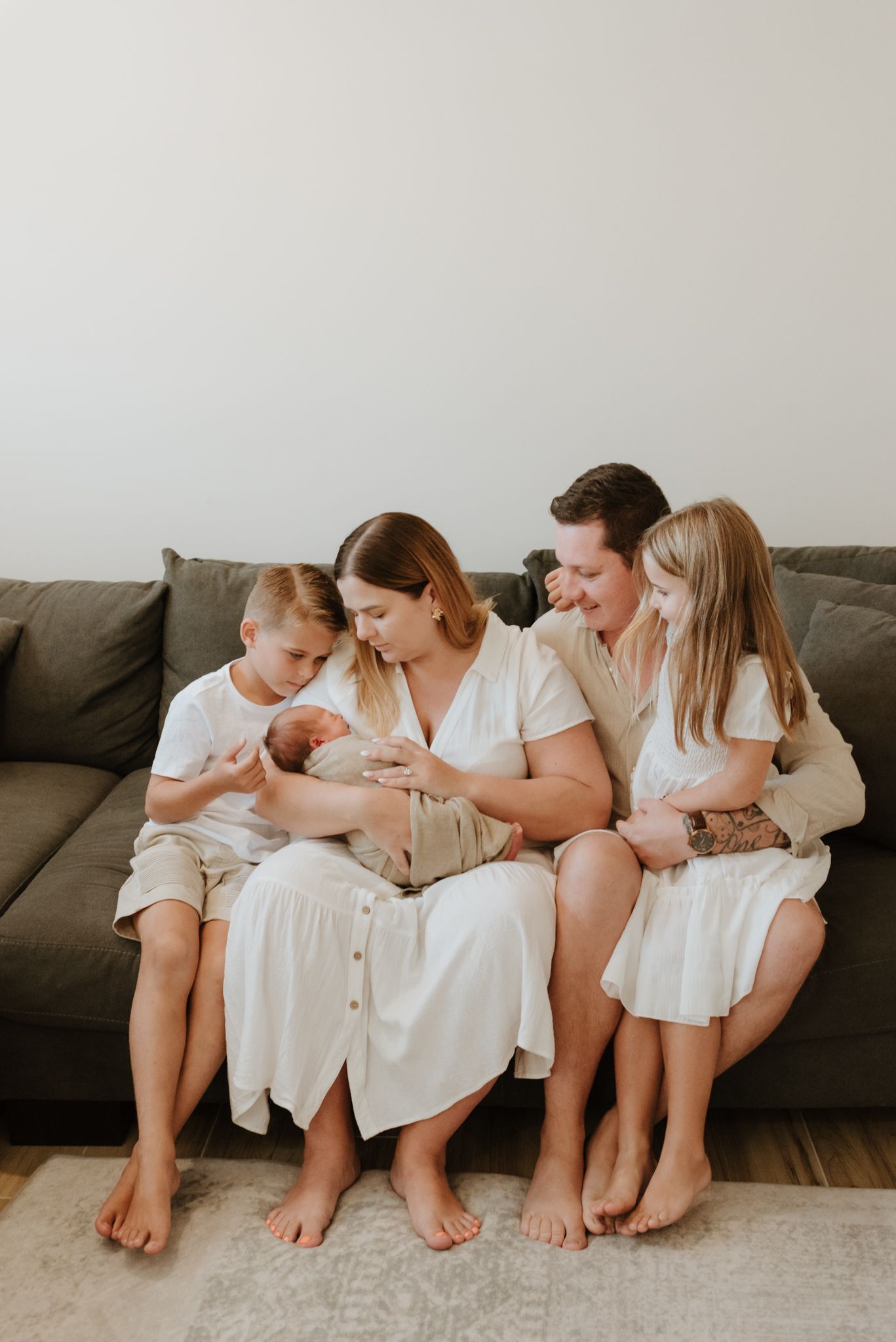 Family sitting on dark couch in casual attire admires their newborn baby together in neutral-toned room.