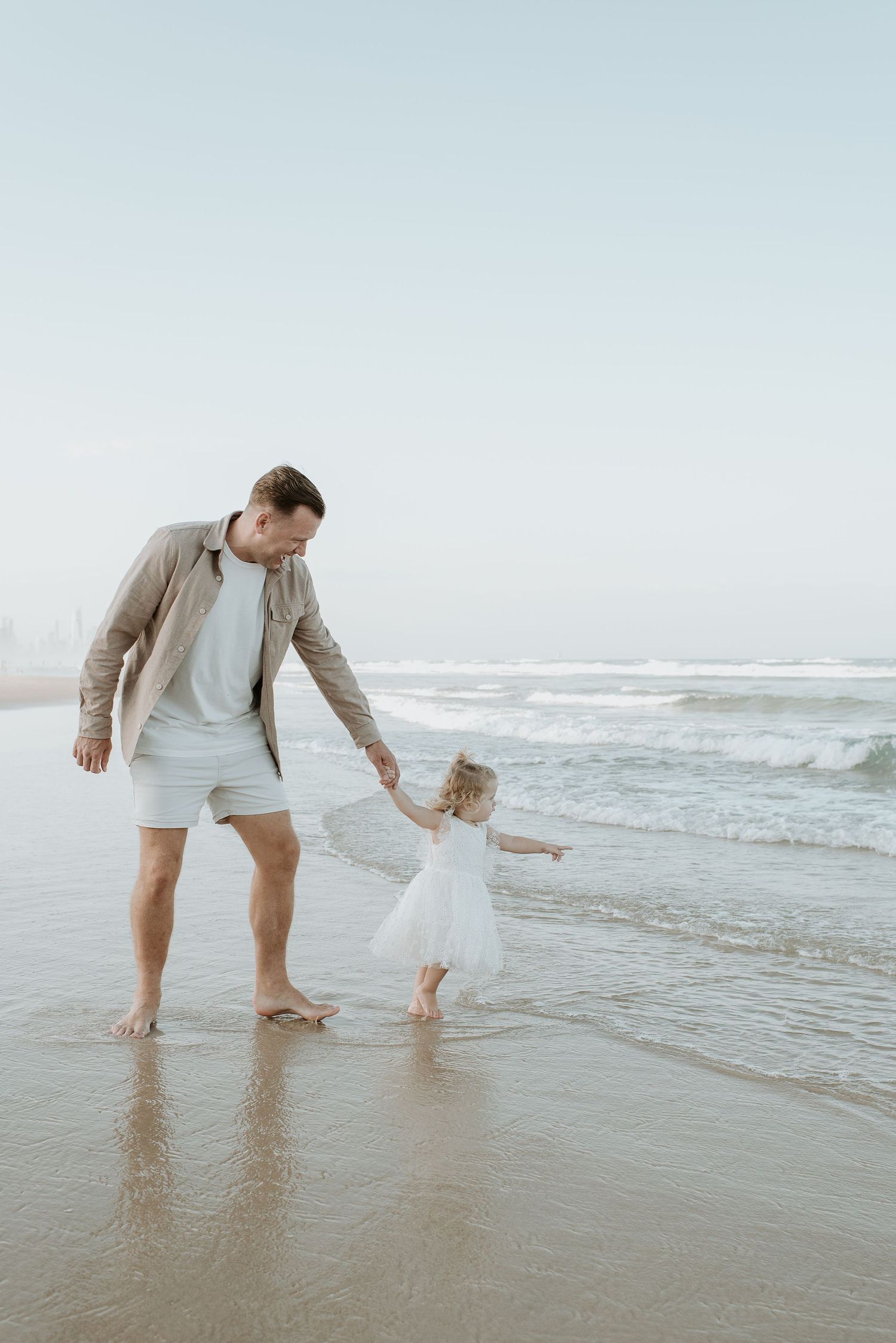 Two figures walking barefoot together on a sandy beach by the ocean waves at sunset.