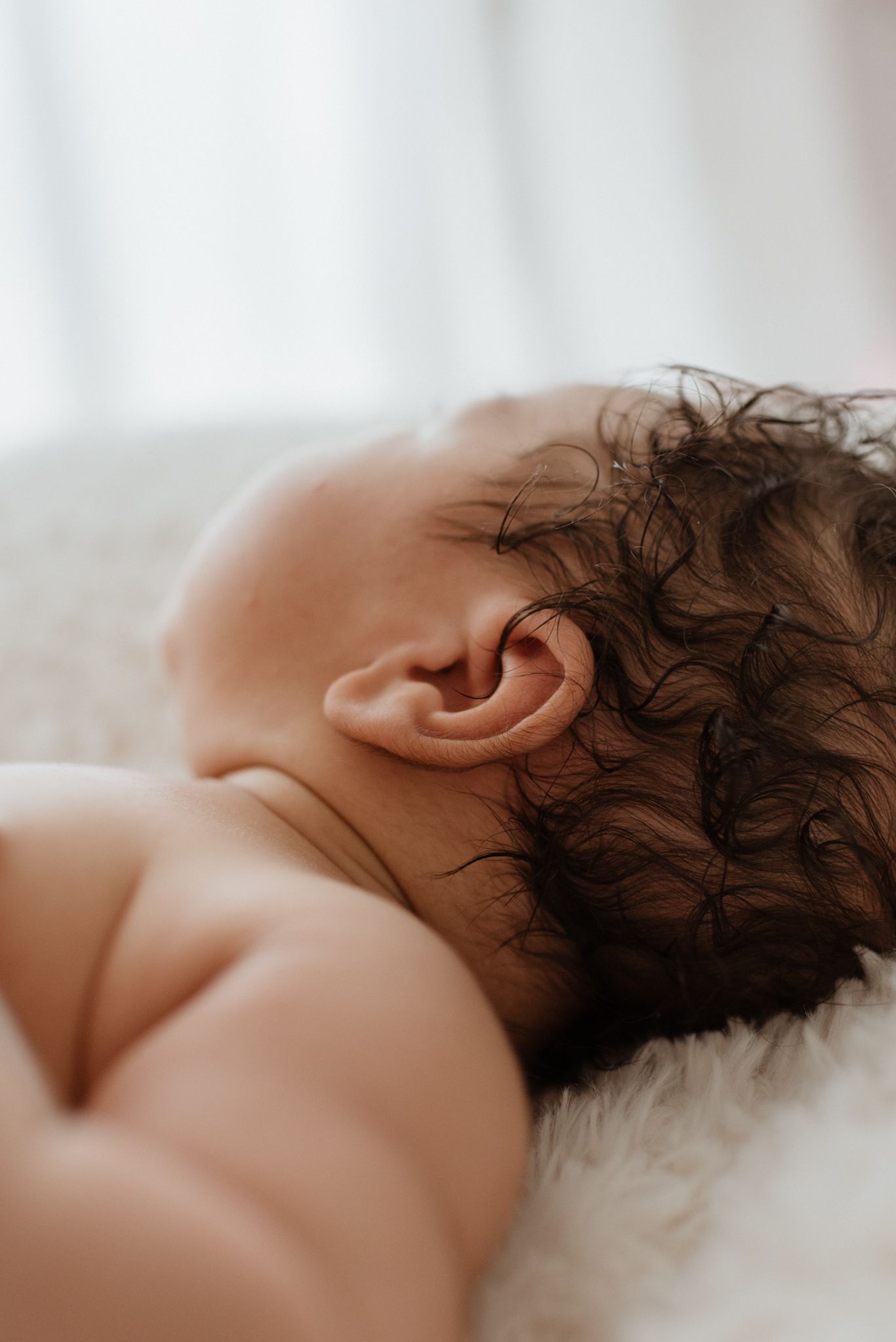 Close-up detail of baby's ear and dark curly hair against white background.