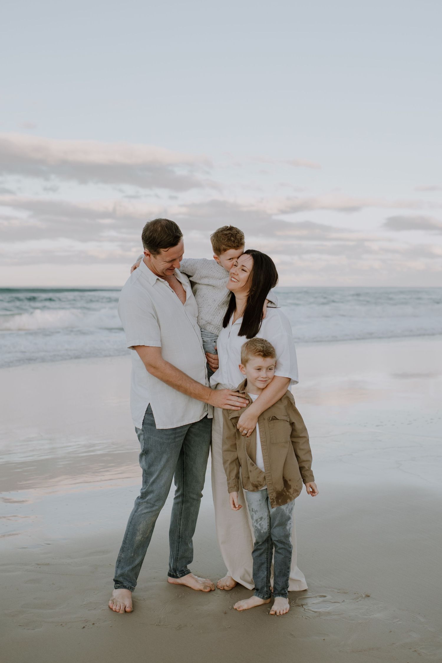 A family of four stands barefoot together on a cloudy beach at sunset sharing an intimate moment.