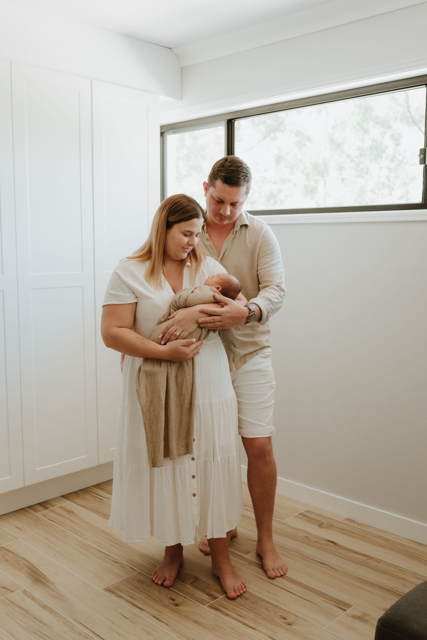 Family wearing light colored clothing stands together on hardwood floor while holding newborn baby in bright room.