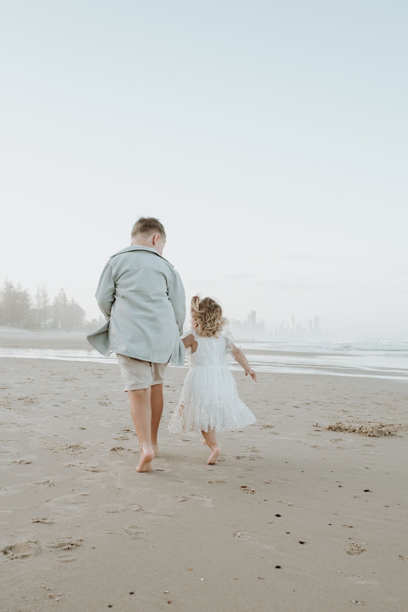 Adult and small child walking barefoot together on a misty beach at sunset.