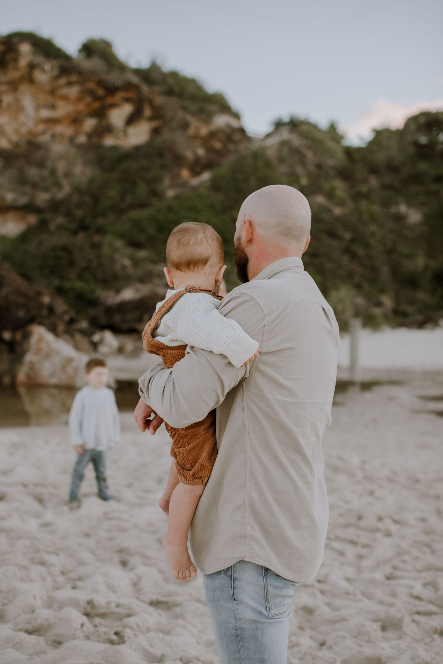Adult holding small child on sandy beach with rocky cliff and another child in background at sunset.