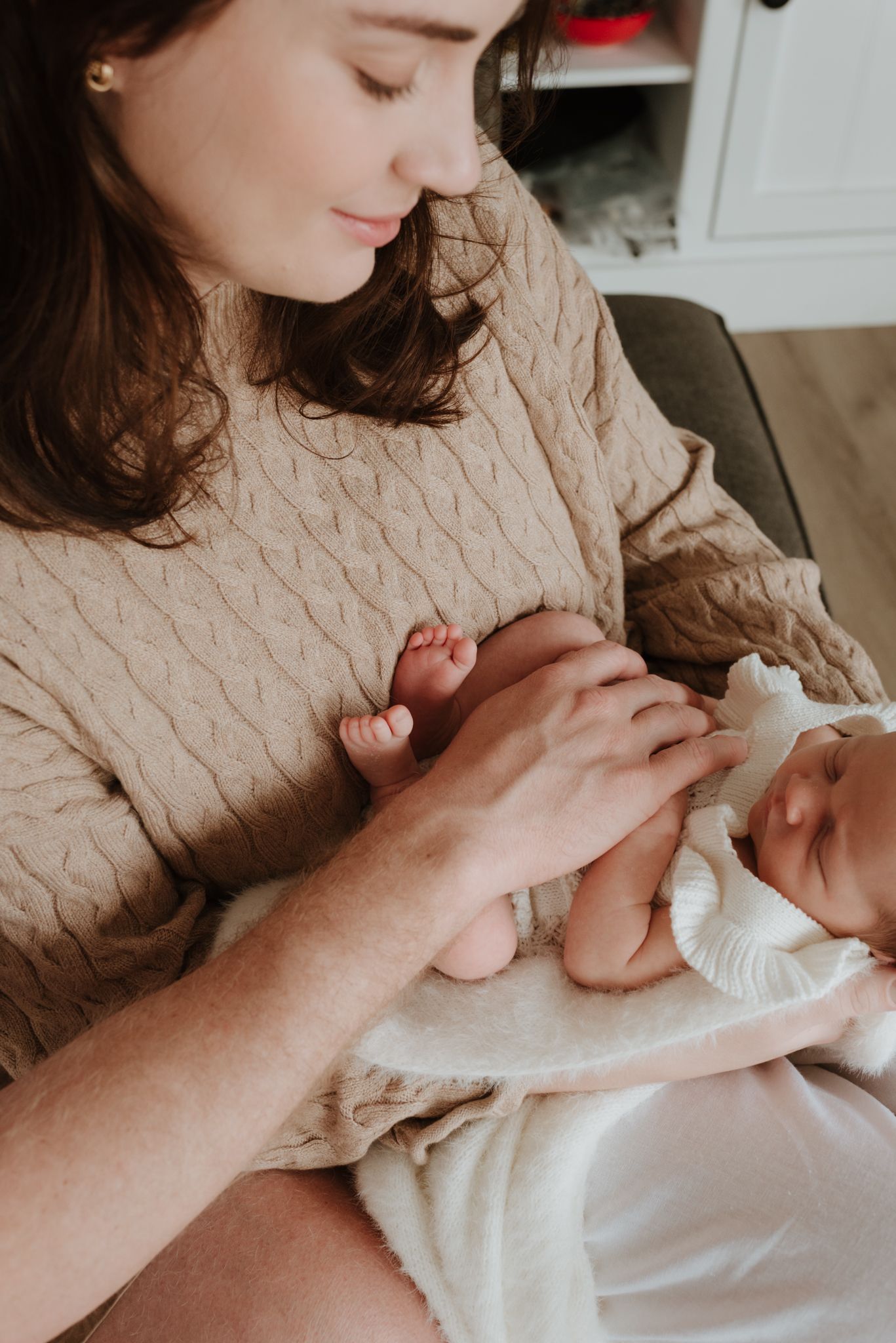 A young mother in a beige cable-knit sweater tenderly holds her sleeping newborn baby wrapped in a white blanket.