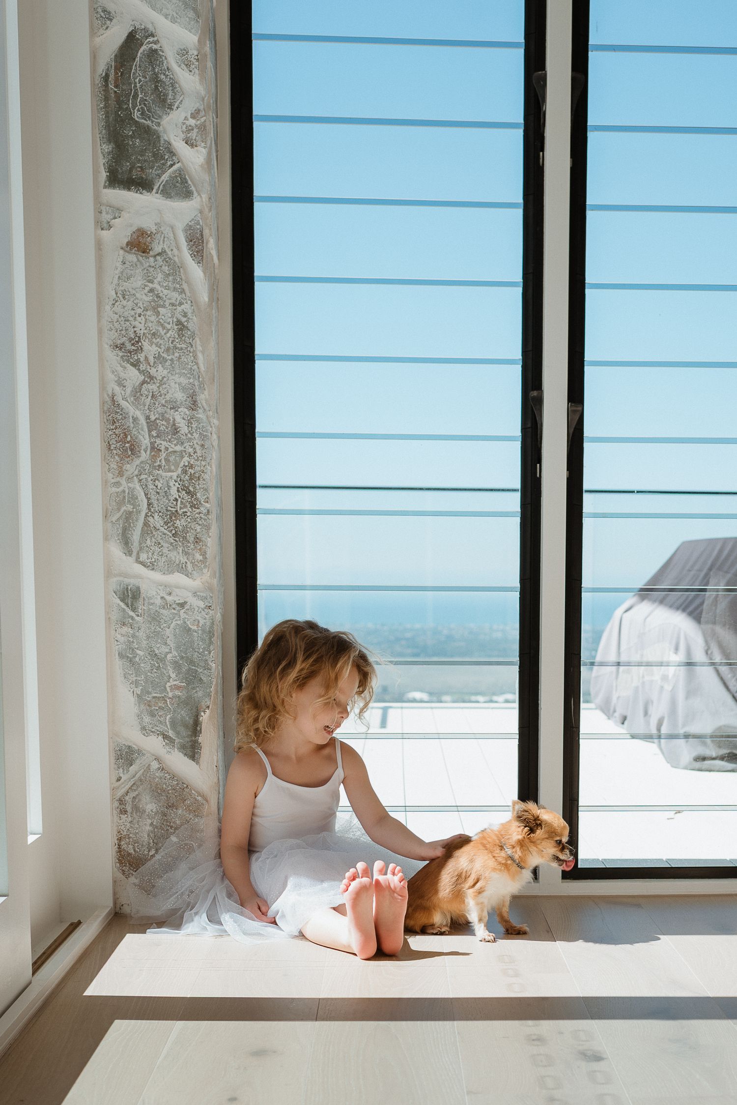 Young girl in white dress sits barefoot by large window with small Pomeranian dog in sunlit modern home.