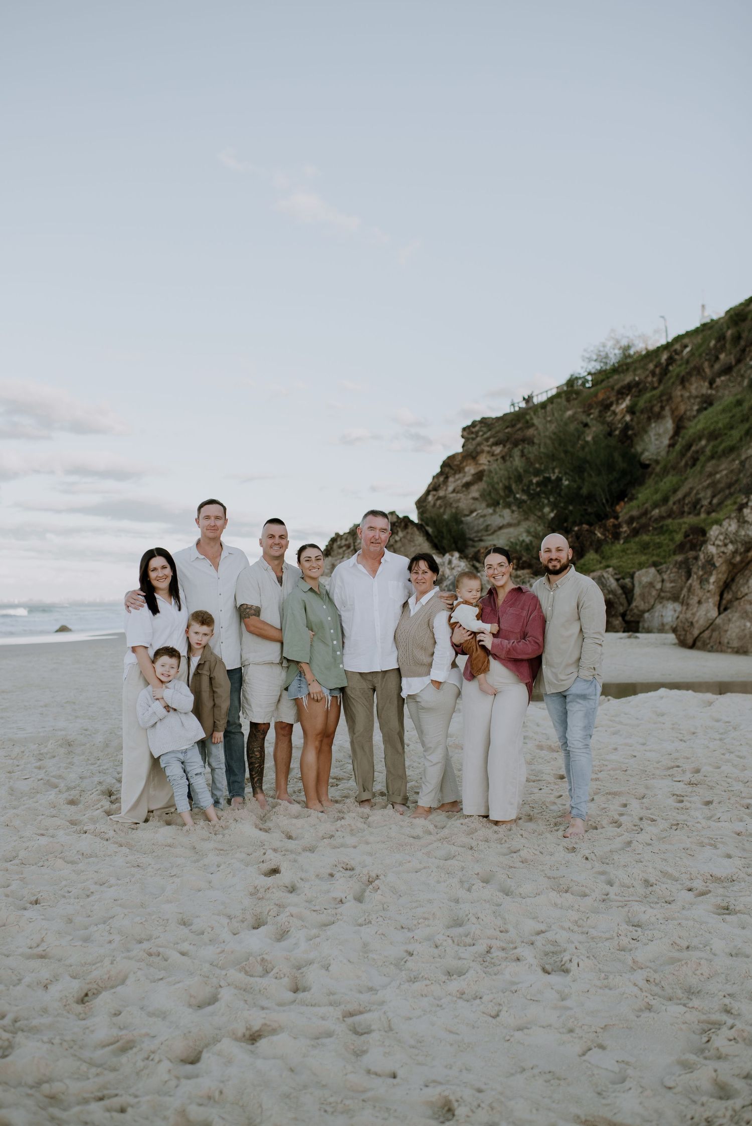 A group poses together on a sandy beach with rocky cliffs and ocean waves in the background on a cloudy day.