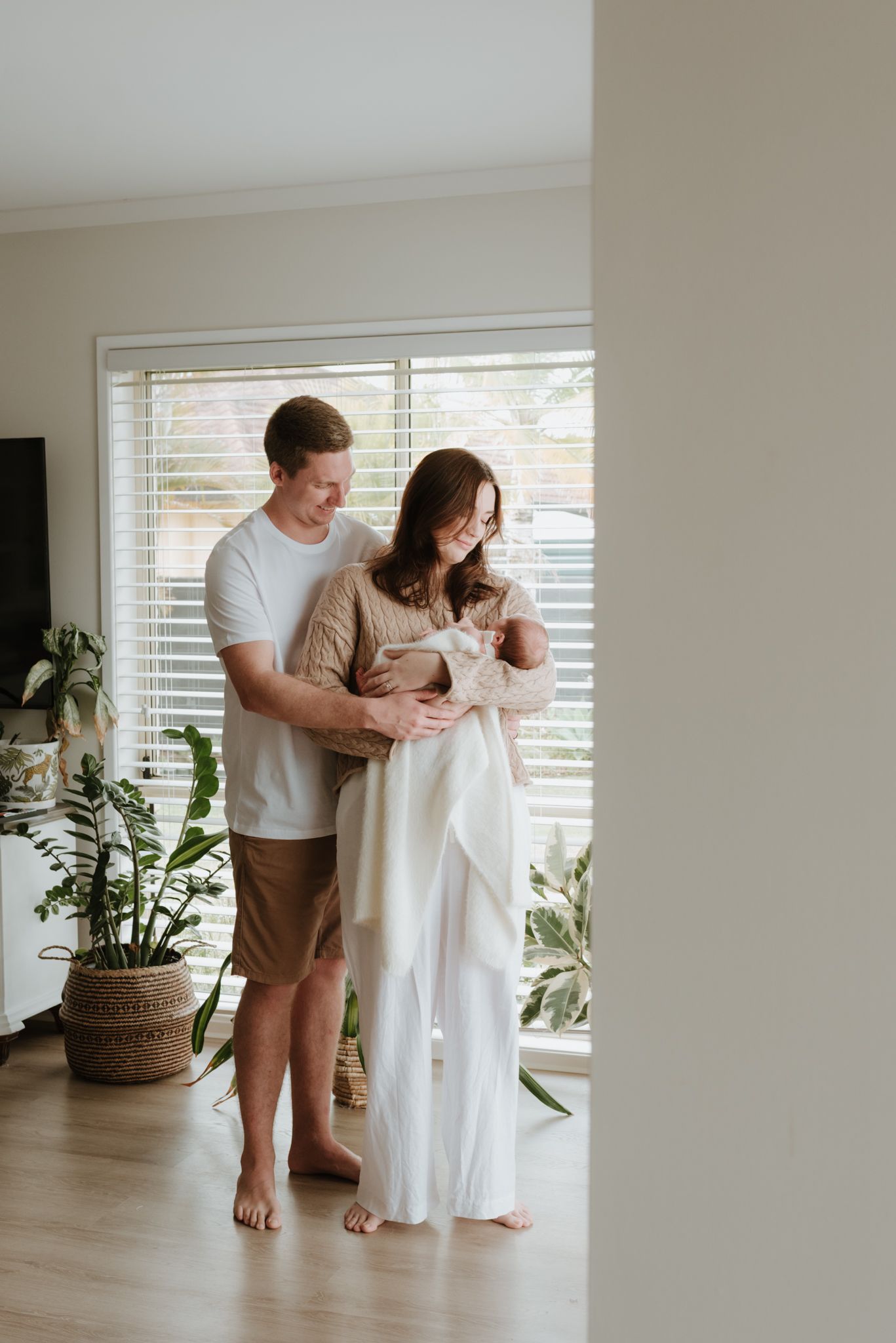 A couple stands indoors holding their newborn baby, wrapped in a white blanket, near a bright window with plants nearby.