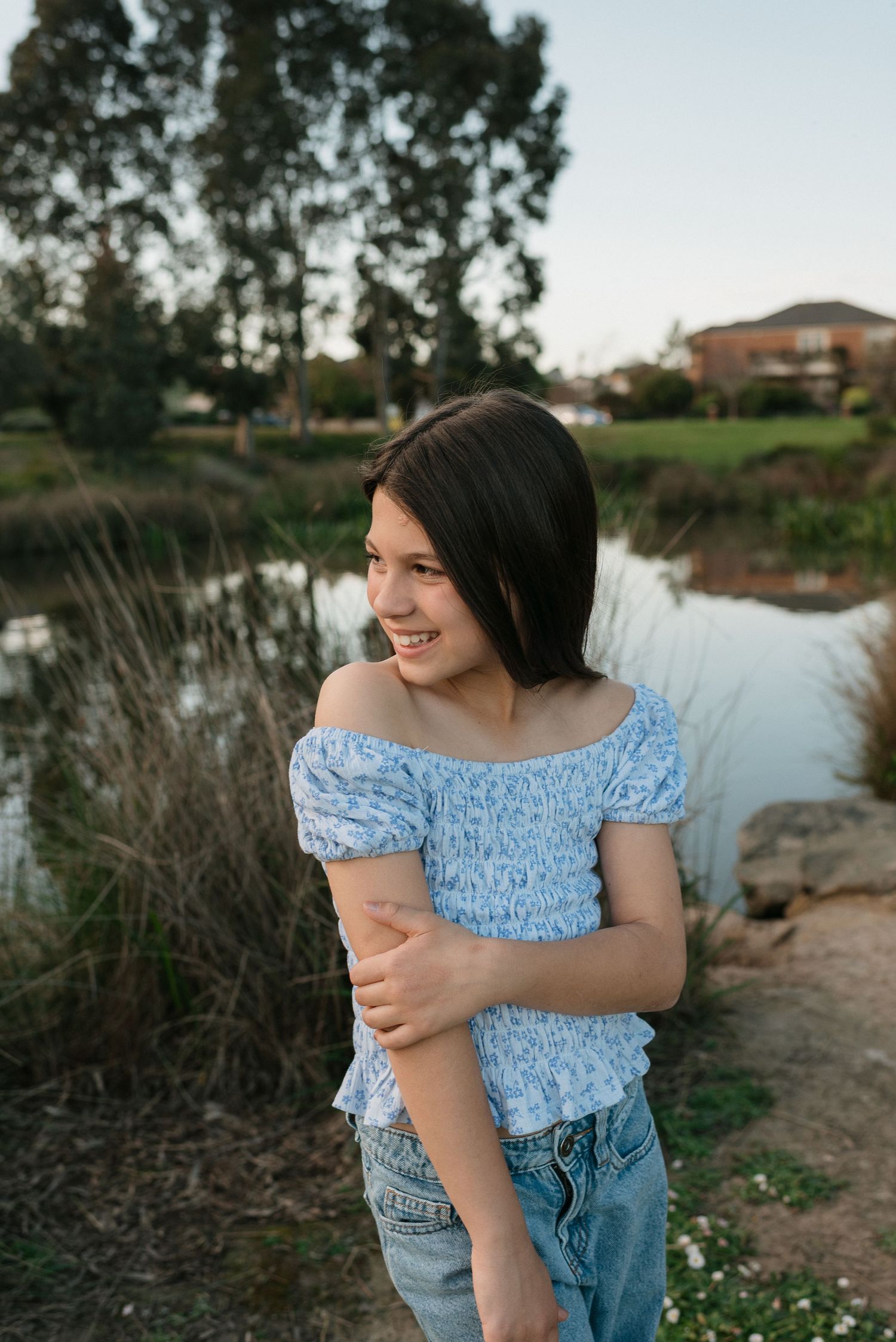 Smiling young woman in blue off-shoulder top and jeans standing near a pond with trees in the background.