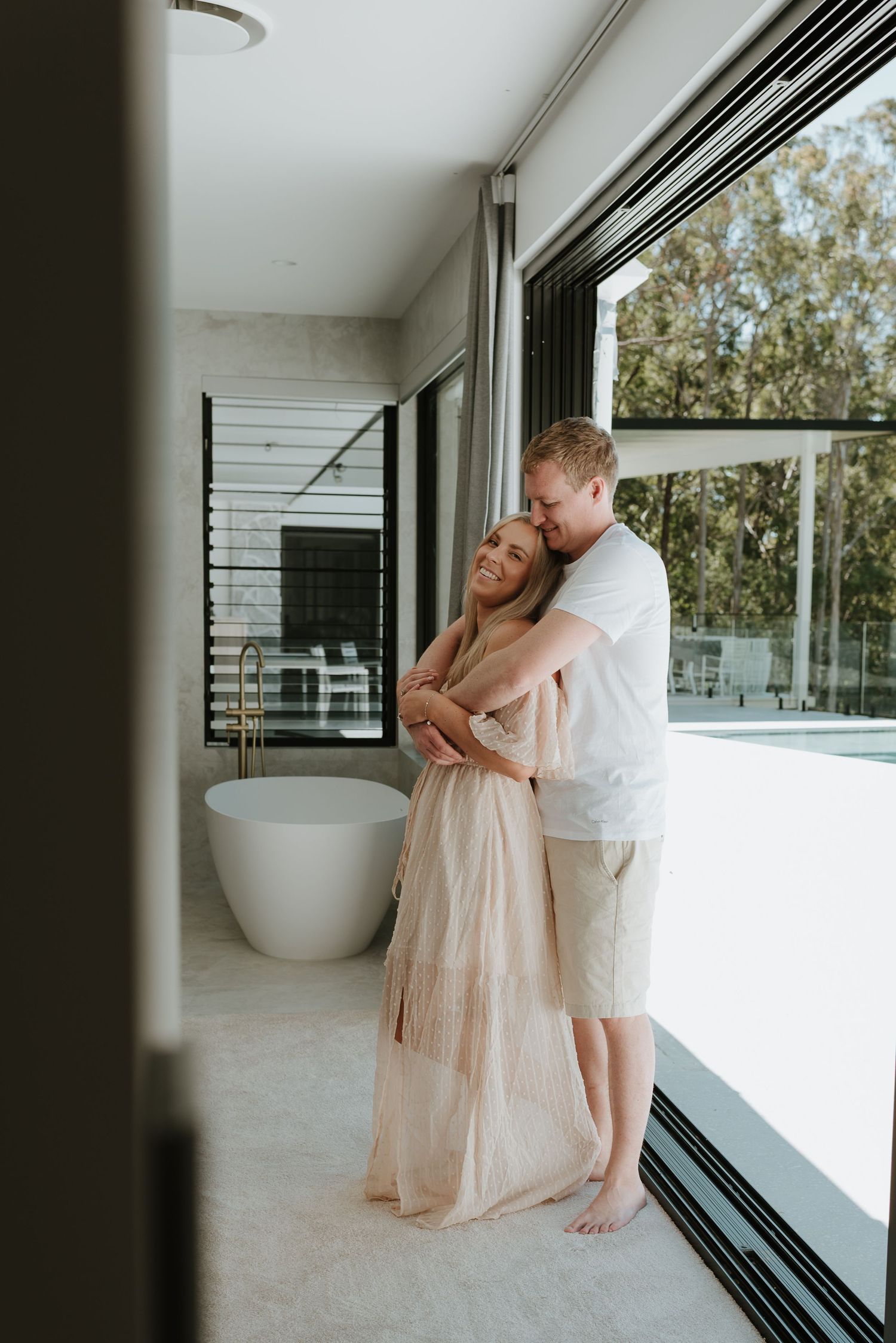 A couple embraces tenderly in a modern bathroom with floor-to-ceiling windows, freestanding tub, and lush greenery outside.