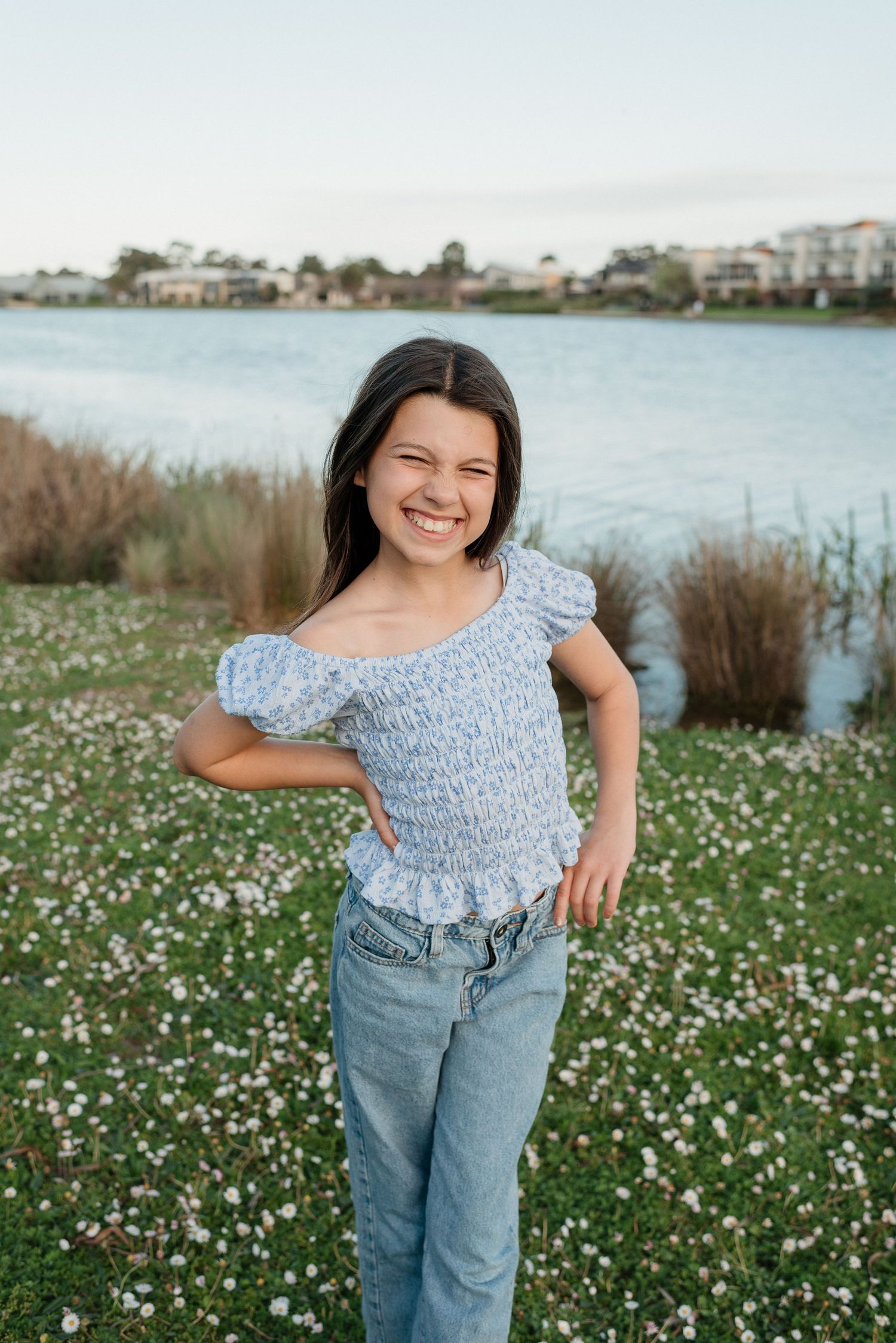 Smiling girl in blue floral top and jeans posing in a wildflower field near a lake.