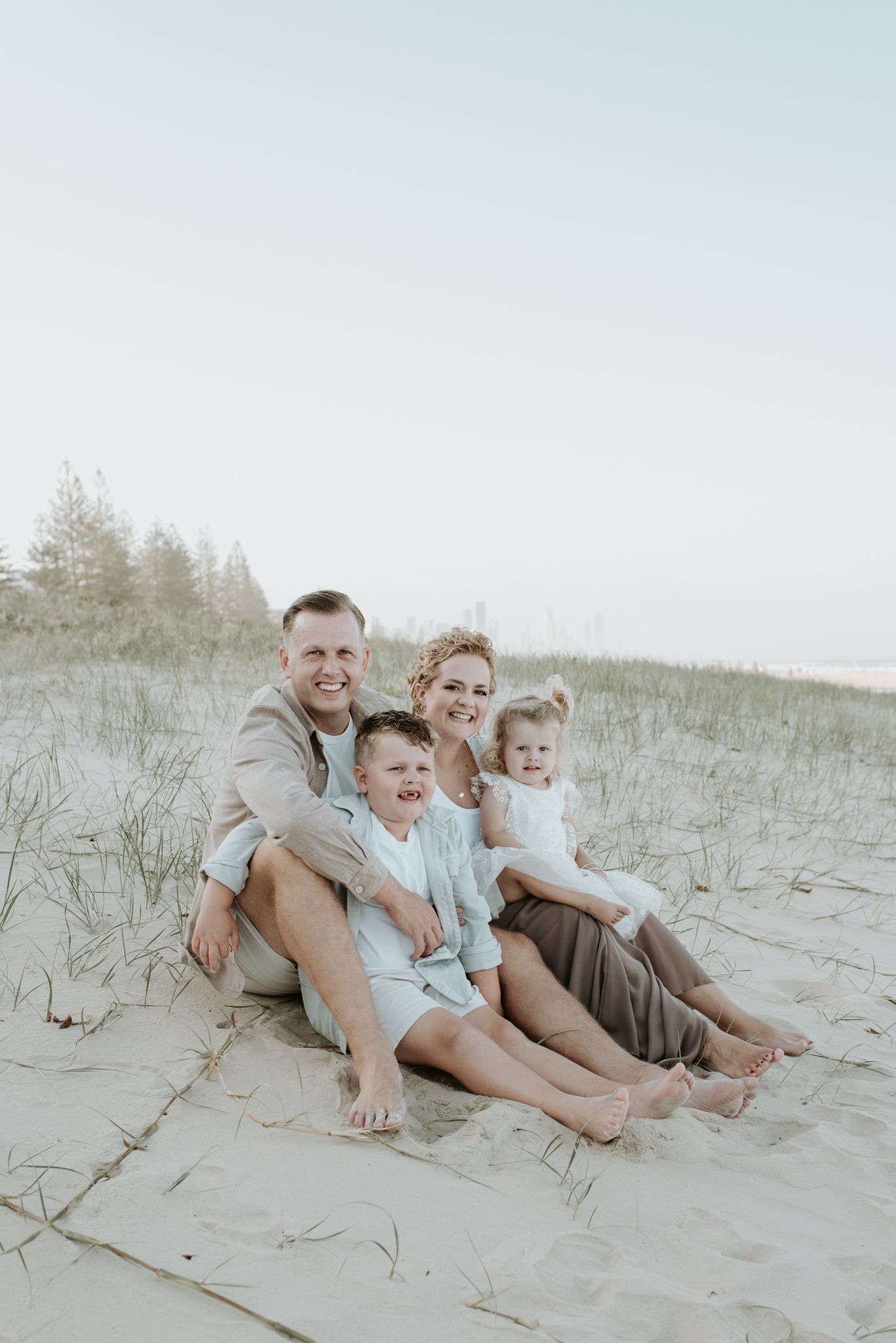 A family of four sits together on a sandy beach dune during a soft, hazy day.