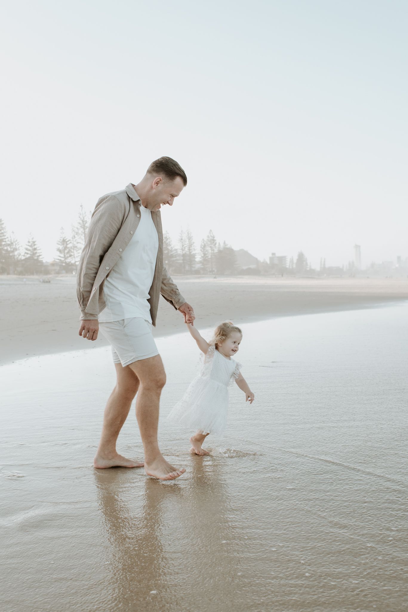 Two people walking barefoot in shallow ocean water at sunset on a misty beach.