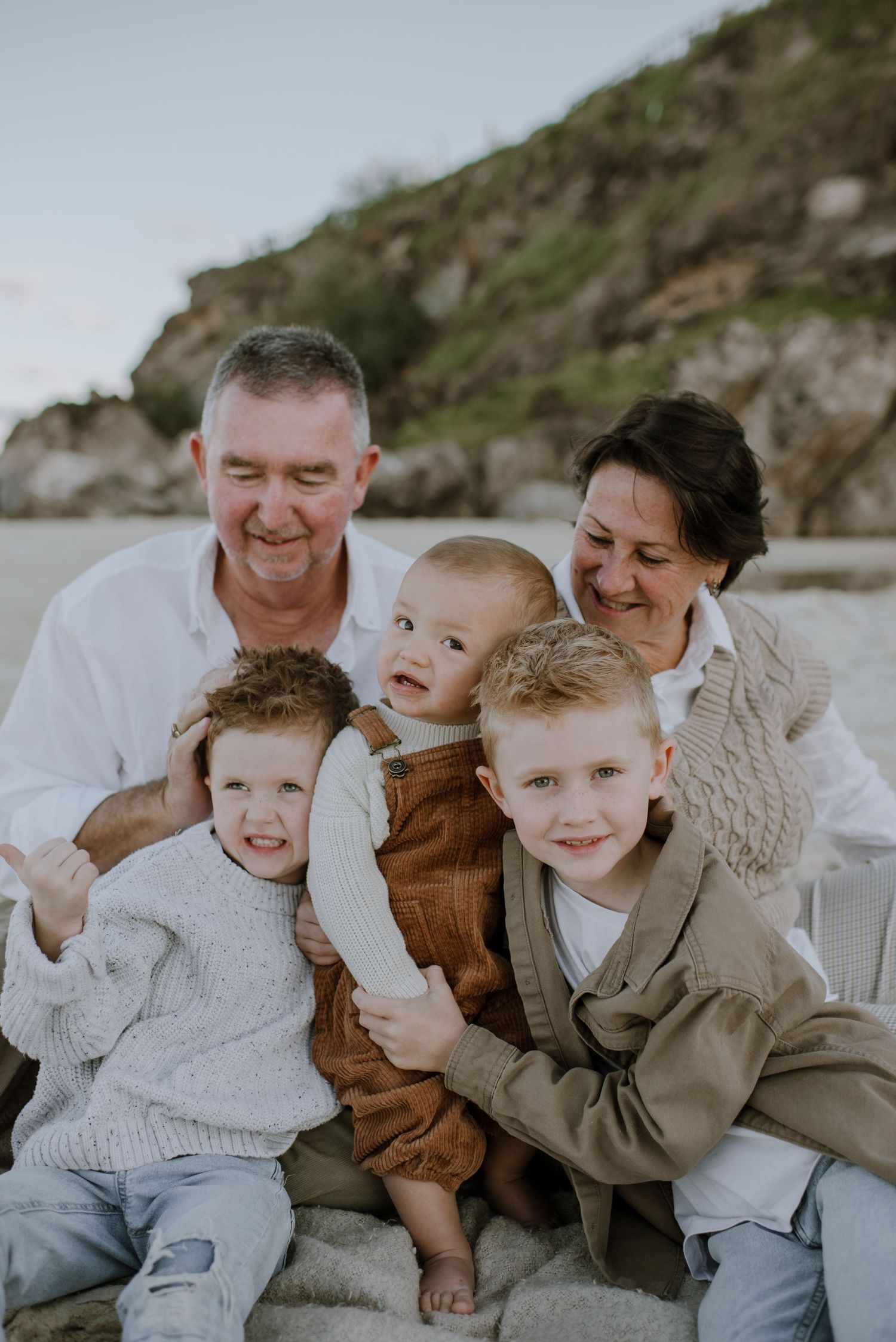A family of five sits together on rocky beach terrain wearing coordinated neutral-colored sweaters and casual attire.
