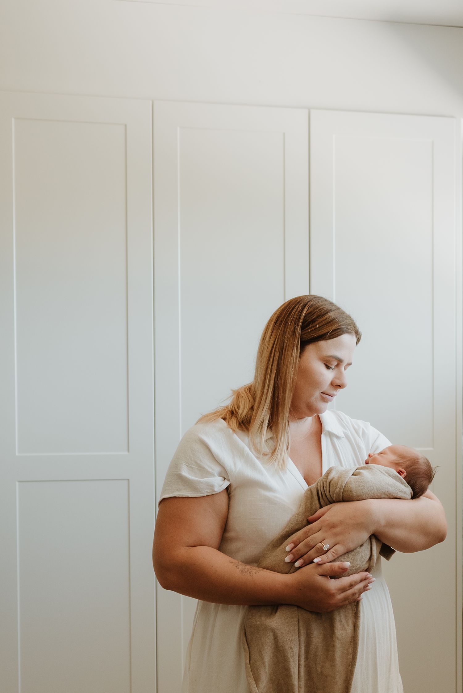 A woman in a white dress holds and looks lovingly at a newborn baby against a light colored wall.