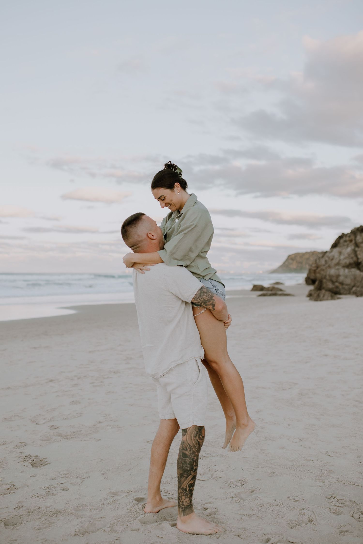 A romantic beach scene with two people embracing on the sand at sunset against a cloudy sky and rocky coastline.
