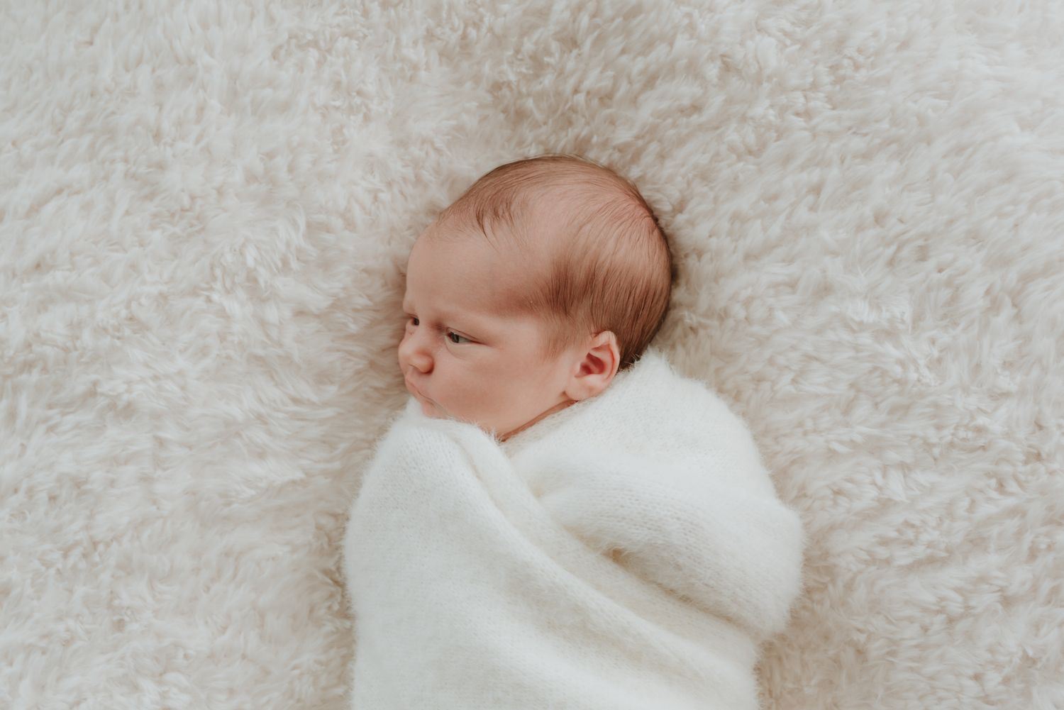 Newborn baby swaddled in white wrap sleeping peacefully on a soft cream fluffy blanket.
