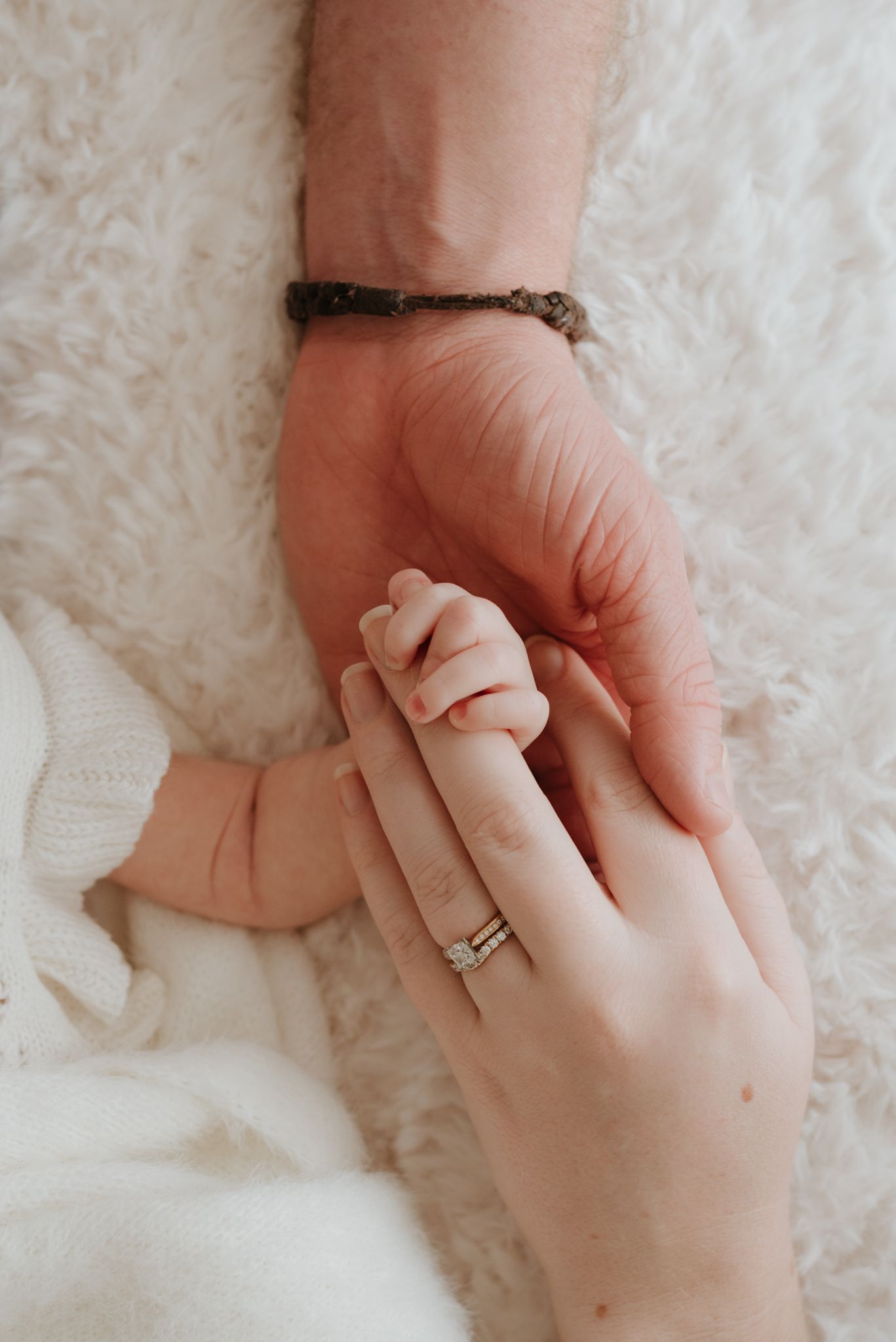A newborn baby grasping an adult's finger, with a wedding ring visible, on a soft white blanket.