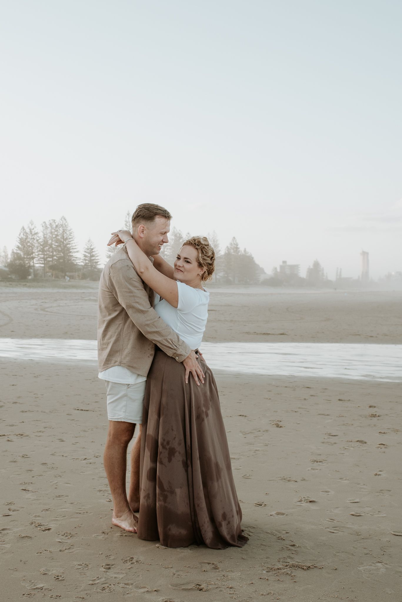 A couple shares a romantic embrace on a misty beach with pine trees silhouetted in the background.