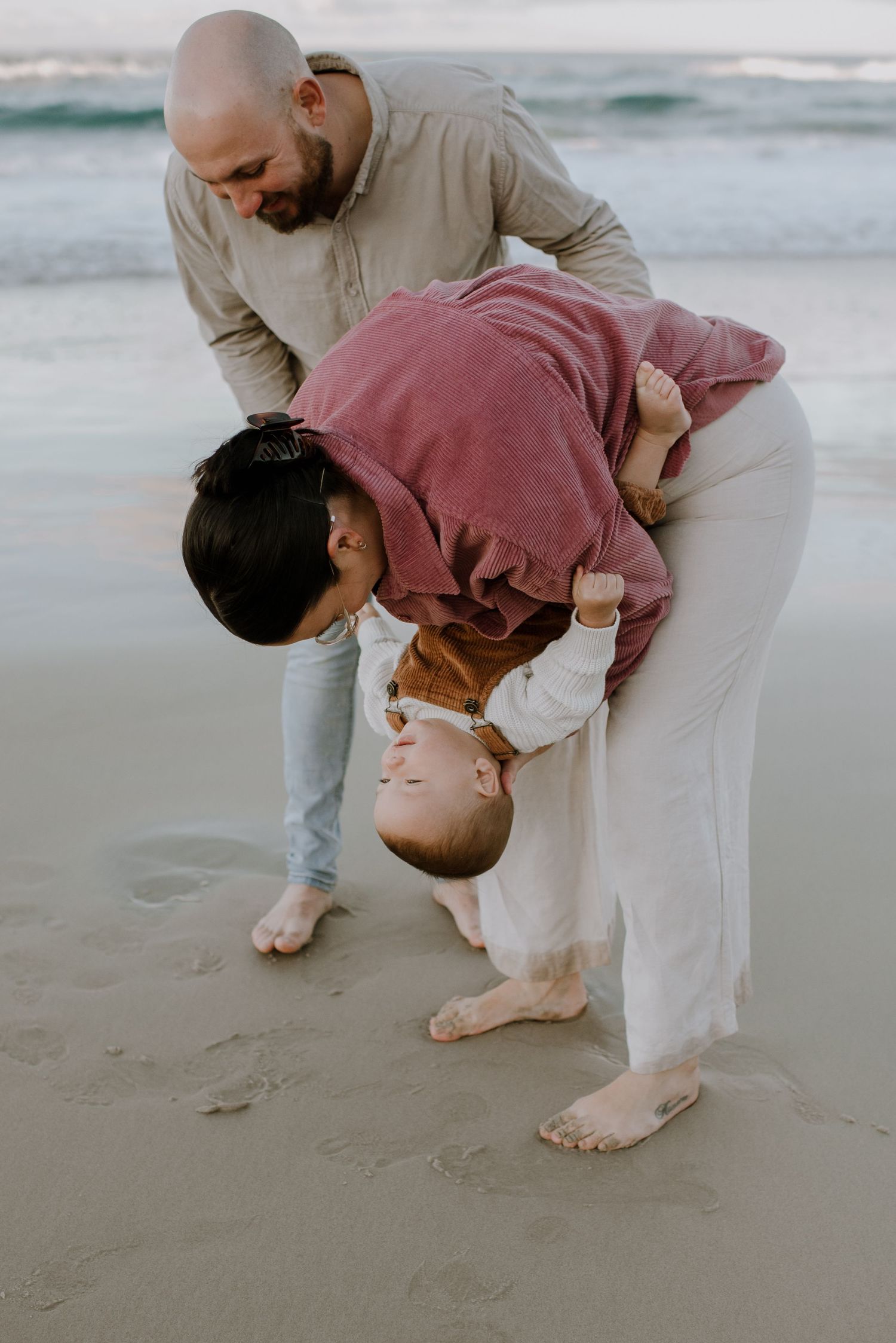 A playful moment on the beach as people embrace and bend over while wearing casual clothing in soft evening light.