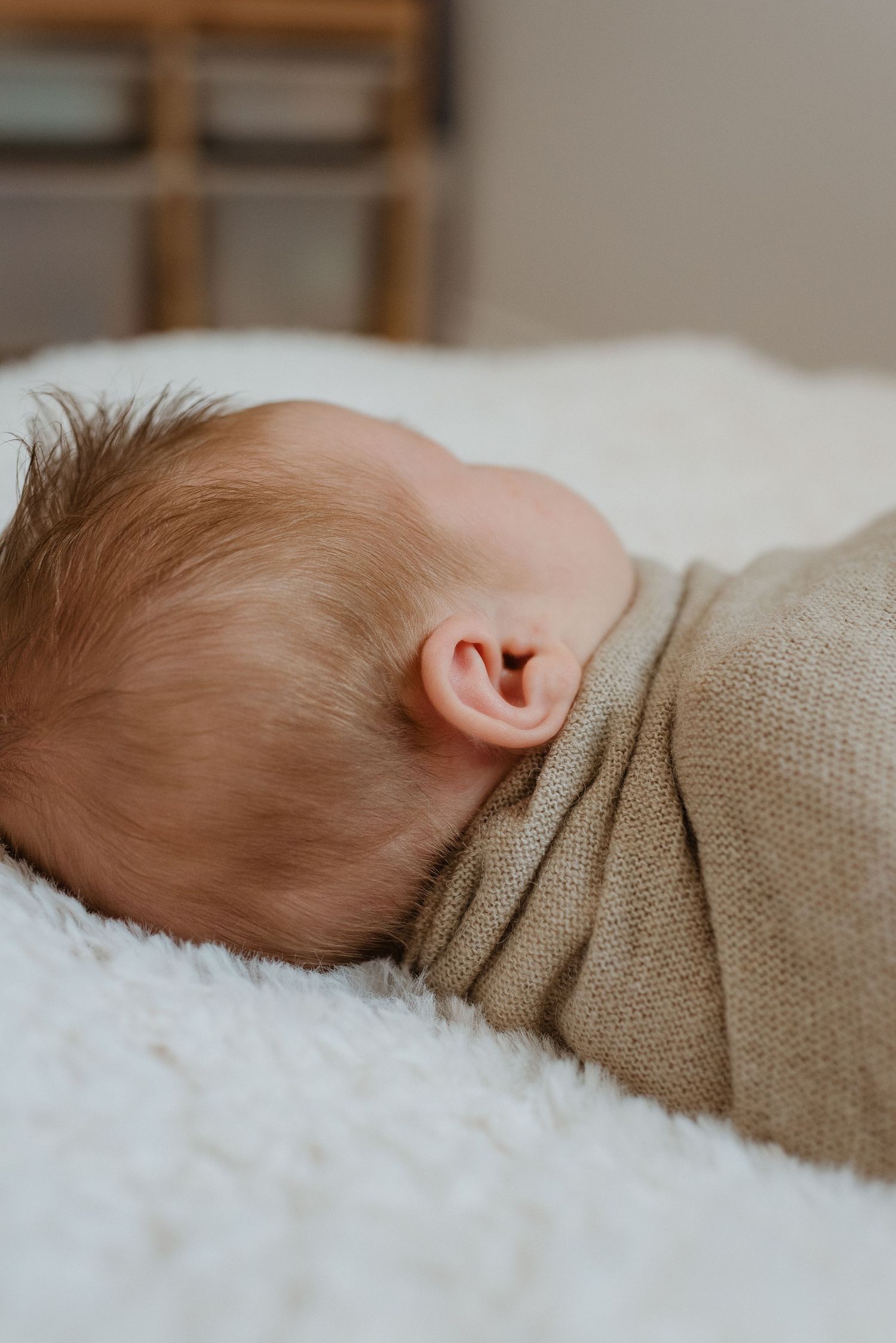 A tiny ear and head of a sleeping baby wrapped in soft beige fabric rests on white fluffy bedding.